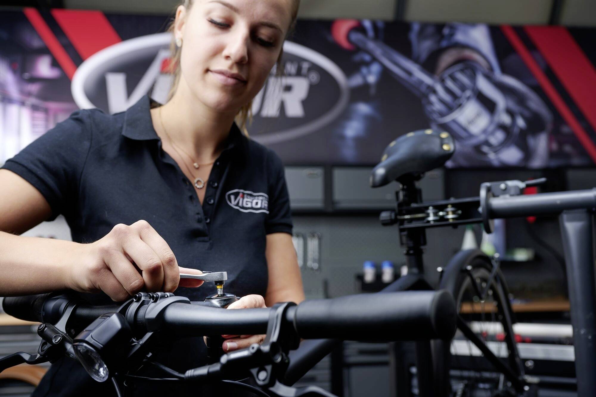 Eine Frau arbeitet an einem Fahrradlenker in einer Werkstatt. Im Hintergrund ist ein großes Schild mit dem Logo 'Vigor' zu sehen.