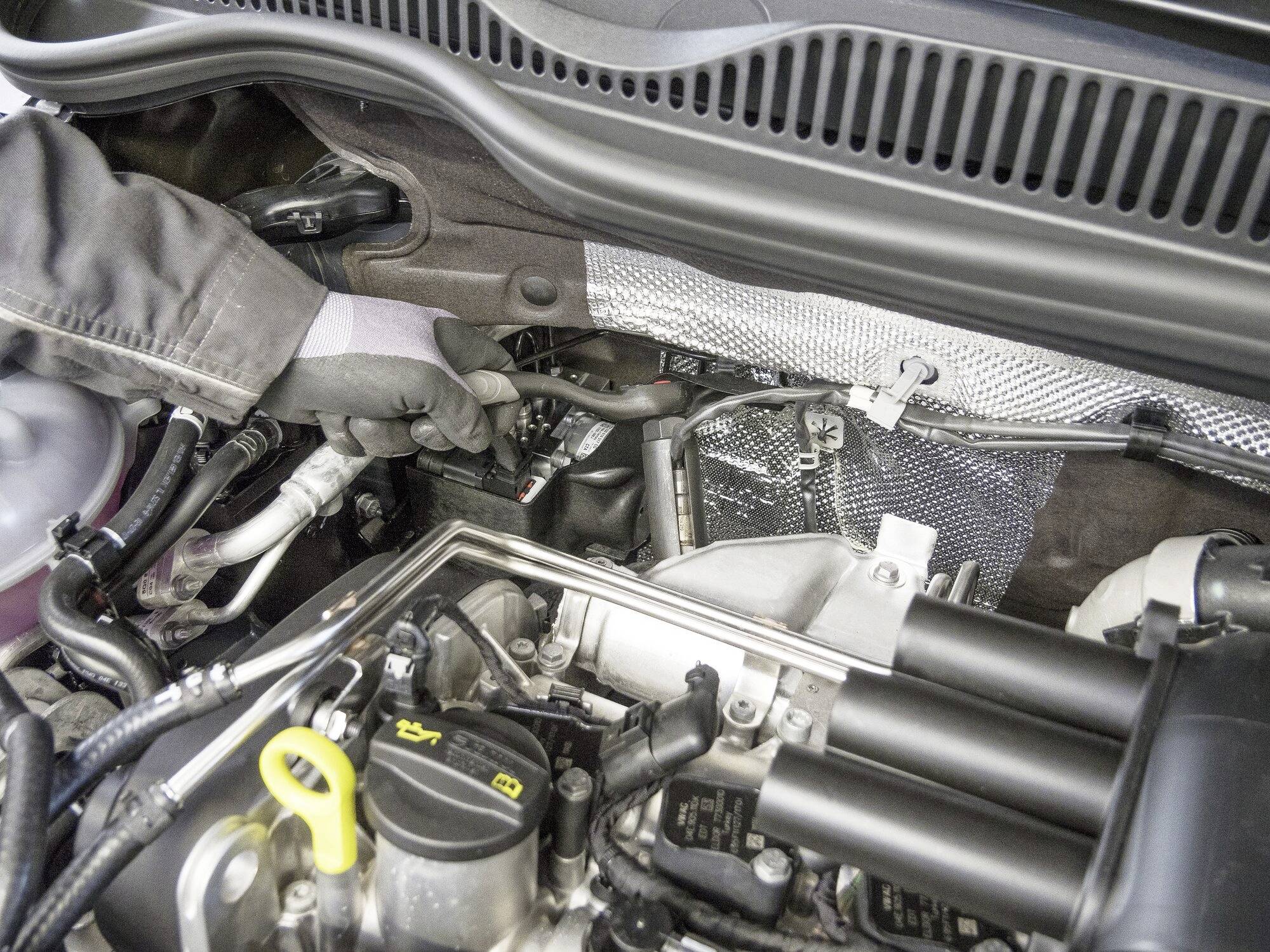 A mechanic is inspecting the engine bay of a car. Various hoses and components are visible. The hand is wearing gloves.