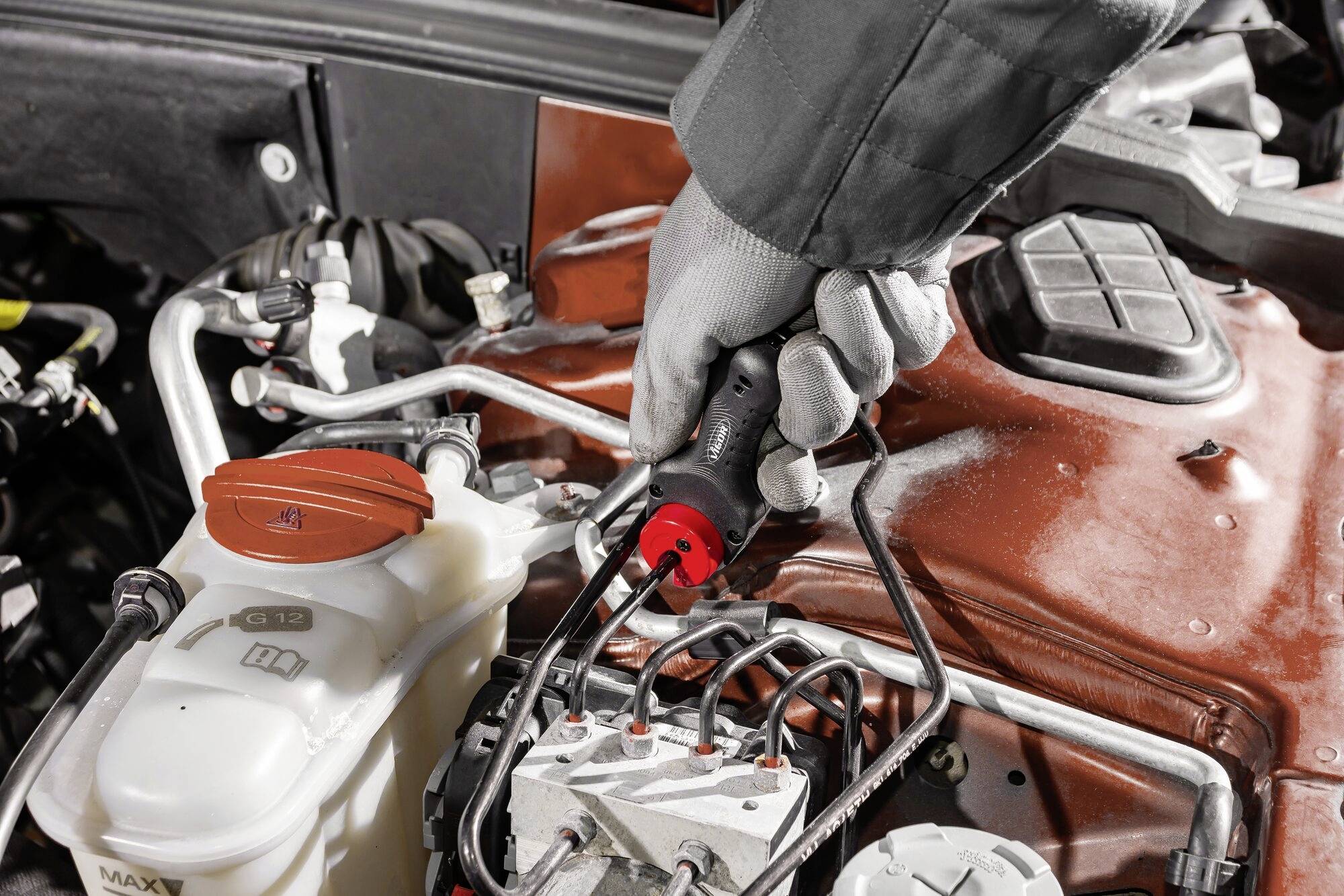 A person wearing a grey glove is using a tool to repair a car's engine. In the foreground, there is a container with liquid.