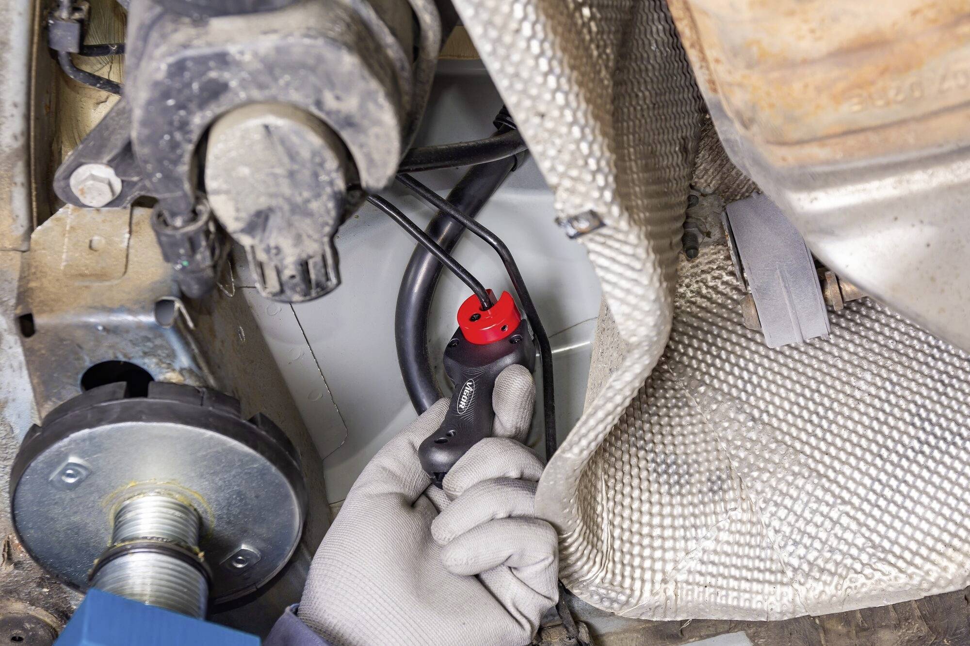 A hand wearing a glove is holding a red diagnostic device near a vehicle part close to the undercarriage and exhaust.