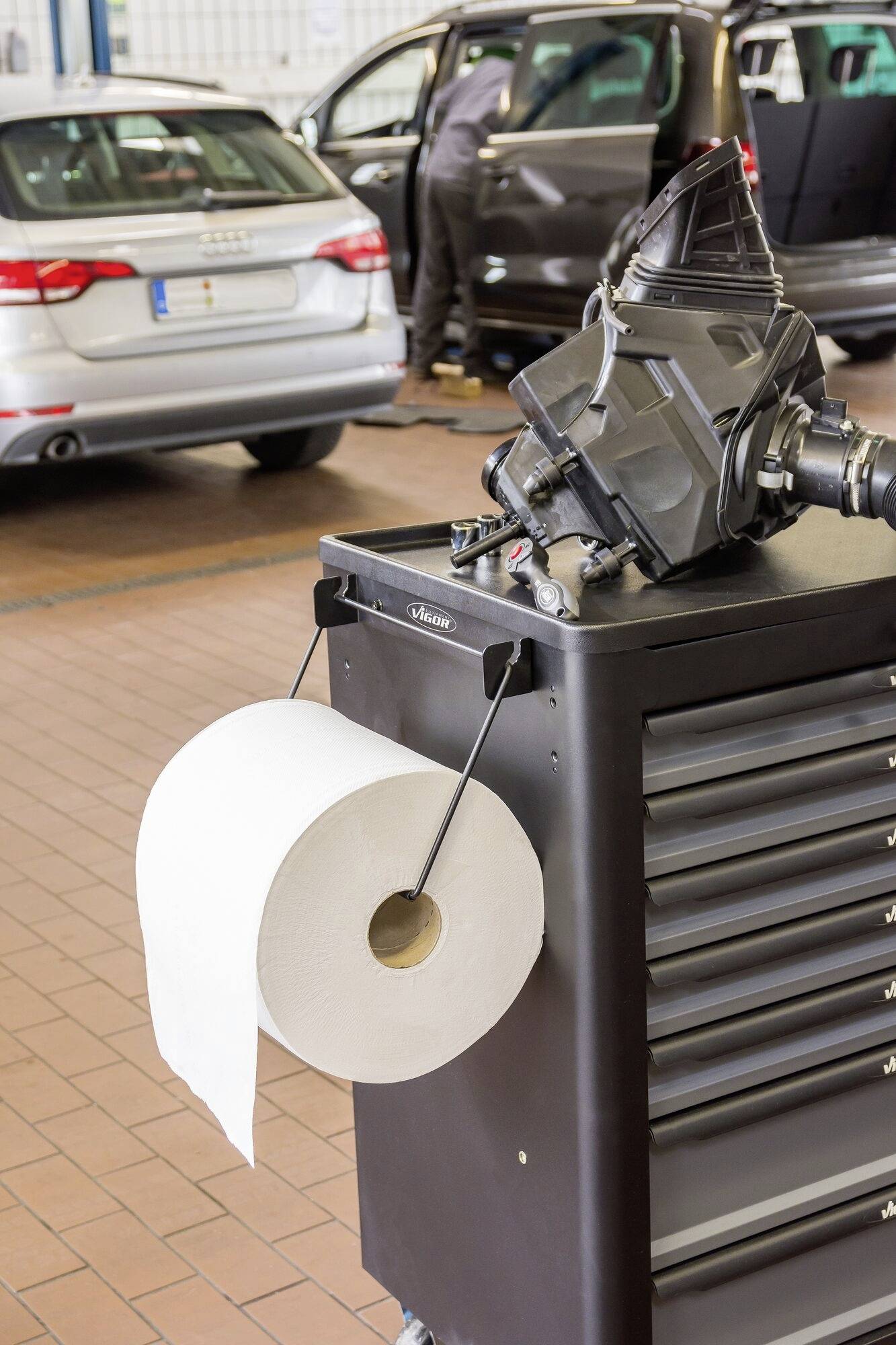 Car Workshop: A trolley with a large roll of paper in the foreground stands next to a mechanic working on a car in the background.