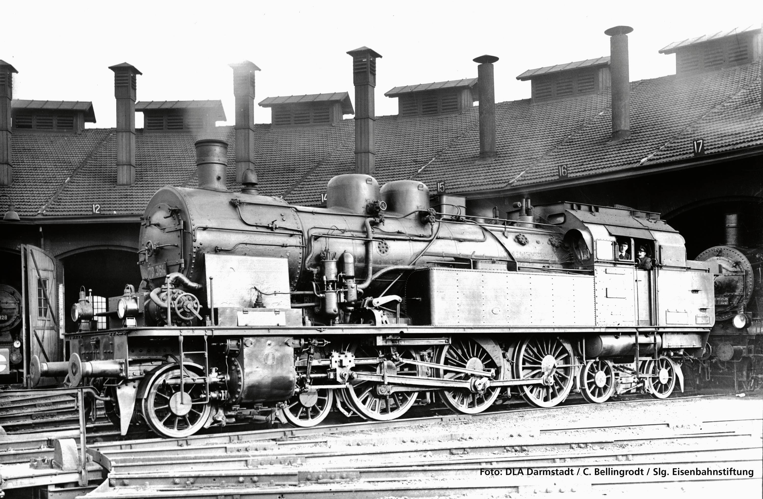 Black and white photograph of a historic steam locomotive in a railway depot, surrounded by trains with buildings in the background.