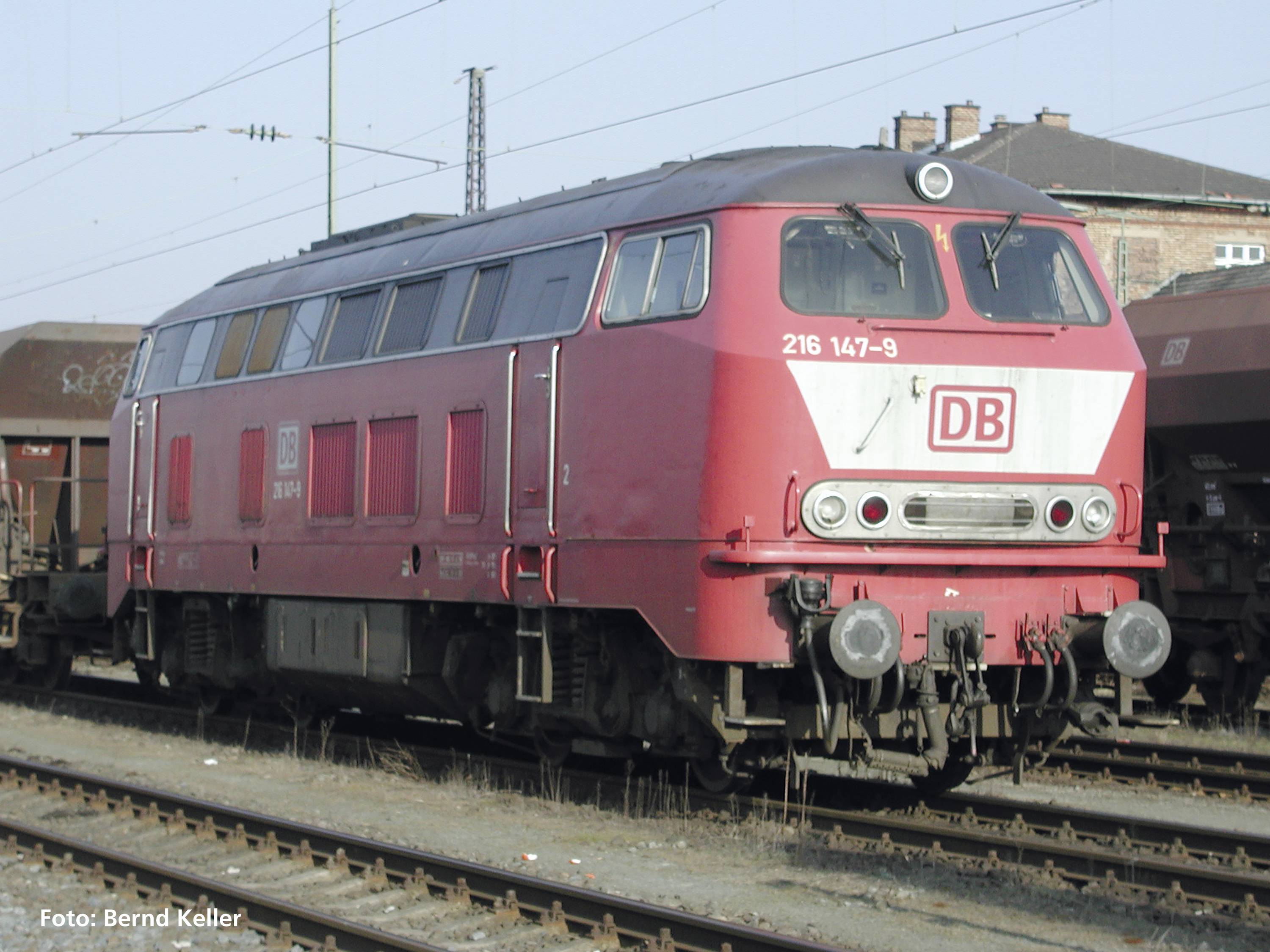A red Deutsche Bahn locomotive is standing on a track. It displays the marking '216 147-9'. Buildings are visible in the background.