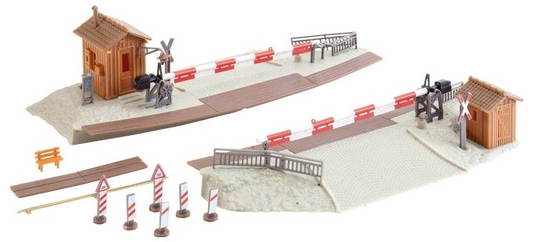 Model railway crossing set with two small wooden huts, barriers, fences, road signs, and tracks on a textured base, illustrating a train crossing scene.