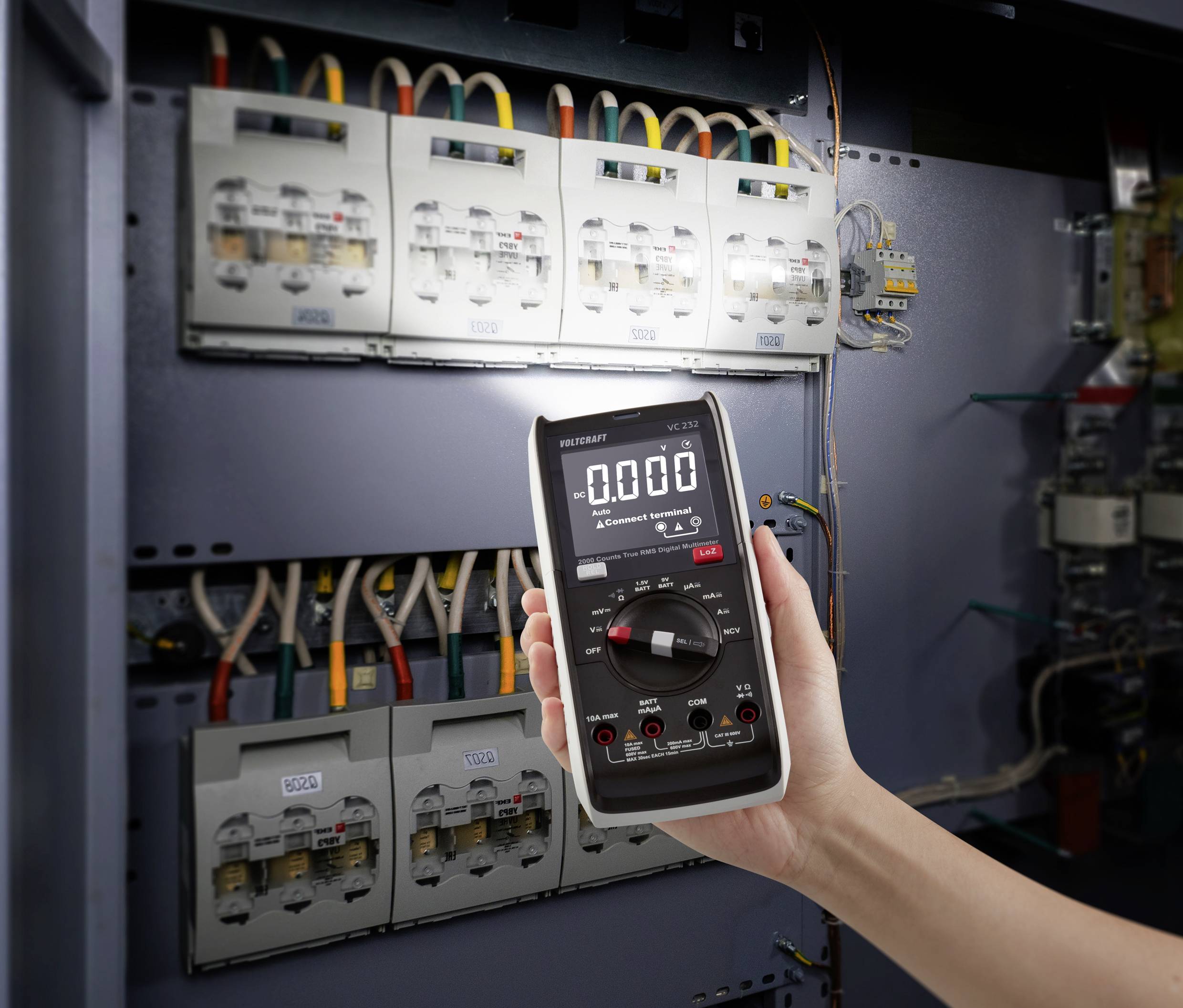 A person is holding a digital multimeter in front of an open electrical cabinet with multiple wirings and fuses.
