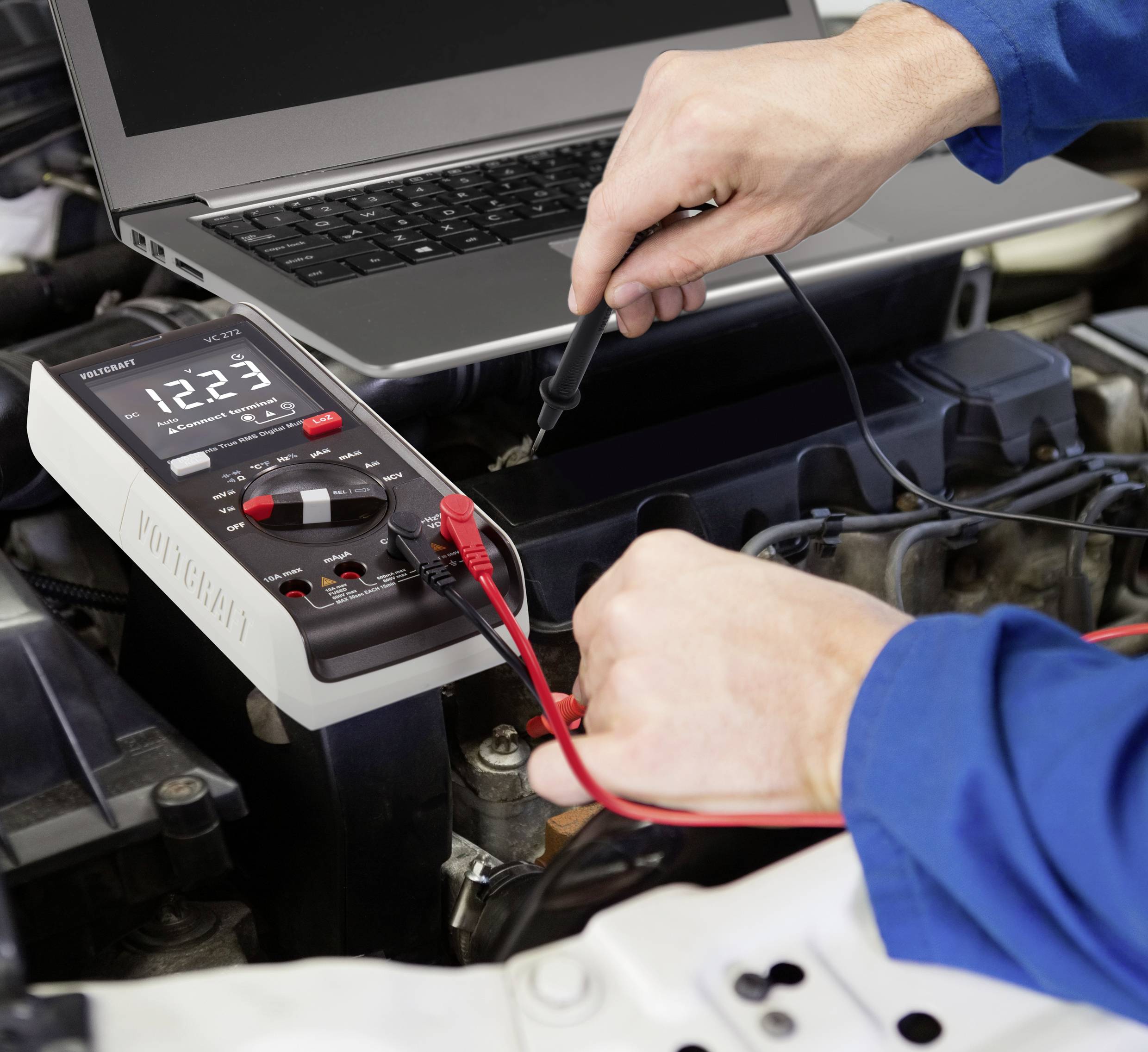 A person is testing the car battery with a multimeter, while a laptop is open nearby.
