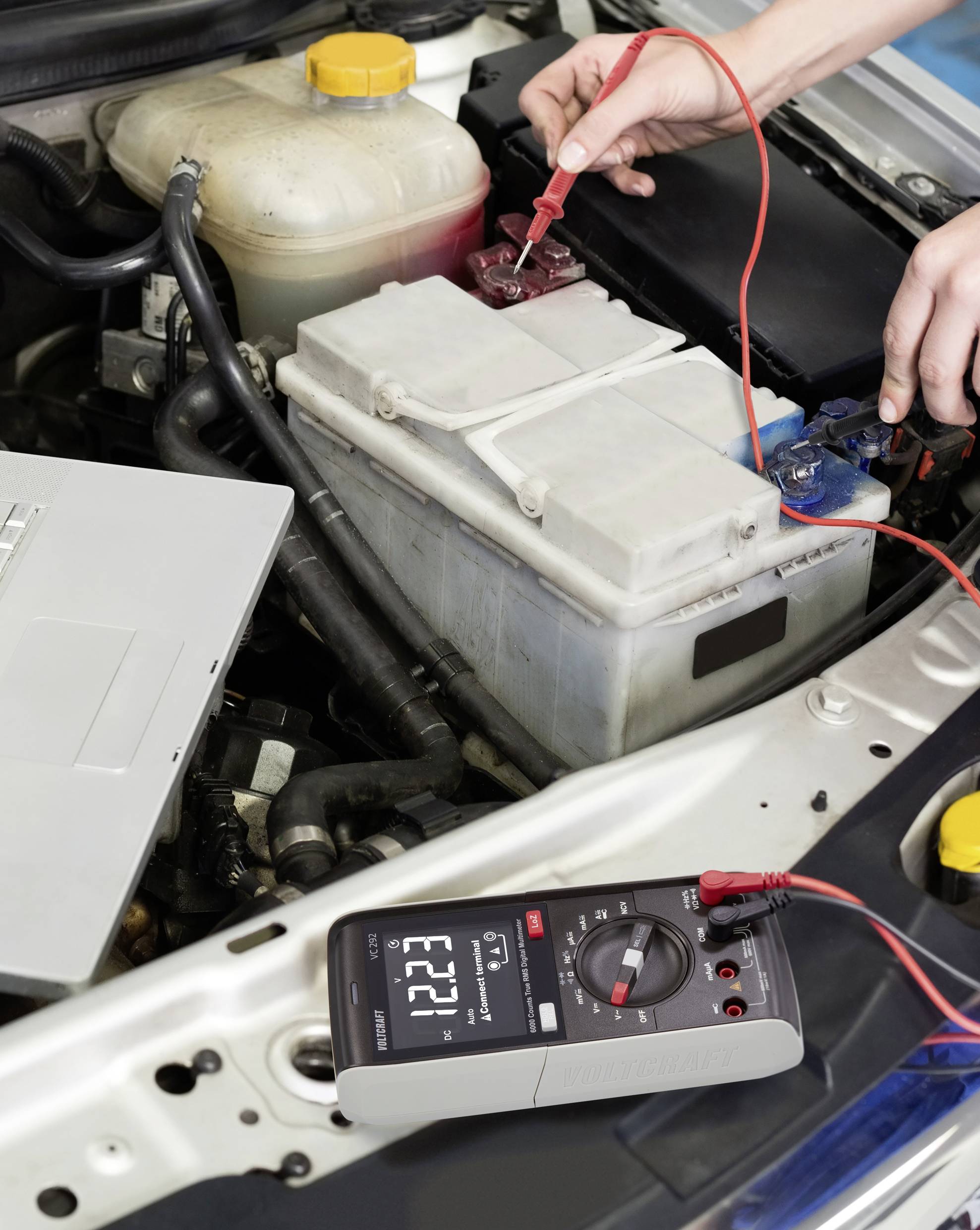 Jemand misst mit einem Multimeter die Spannung einer Autobatterie im Motorraum. Das Display zeigt '12,21' an.
