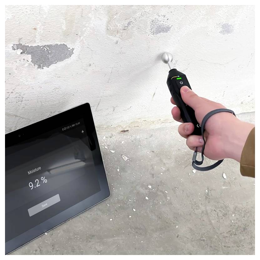 Handheld moisture meter measuring a damp concrete wall while a tablet displays 'Moisture 9.2%'.