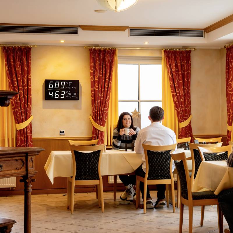 A couple are sitting at a table in a restaurant, surrounded by yellow curtains. A display on the wall shows 68.9°C and 46.3% relative humidity.