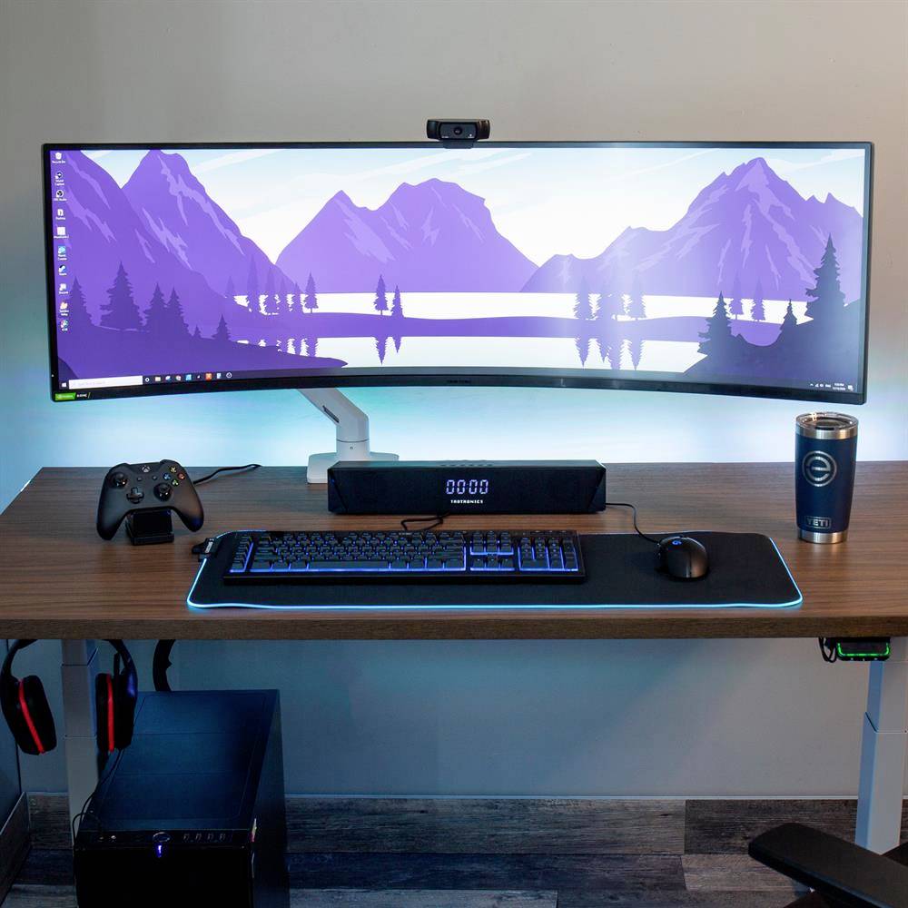 A modern desk with a curved monitor displaying a landscape of mountains and a lake. Controller, speakers, and a mug beside the keyboard.