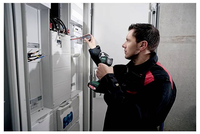 A technician in uniform uses a screwdriver and a drill to work on electrical panels in a control room.
