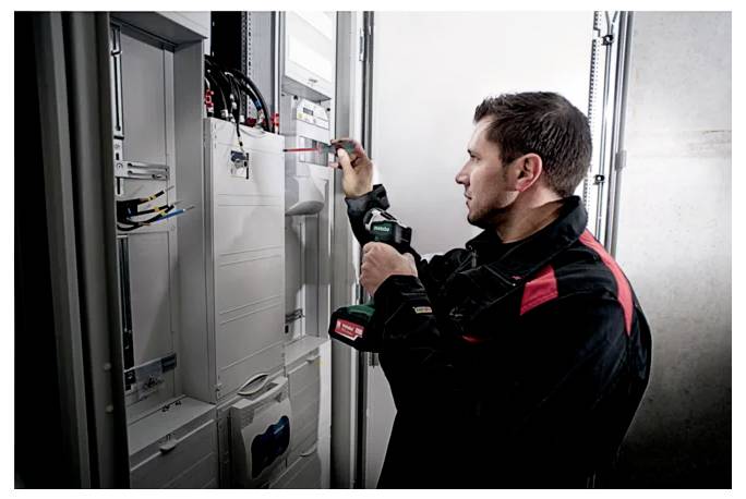 A technician in a black and red uniform uses a drill to work on an electrical panel in a utility room, indicating maintenance activity.
