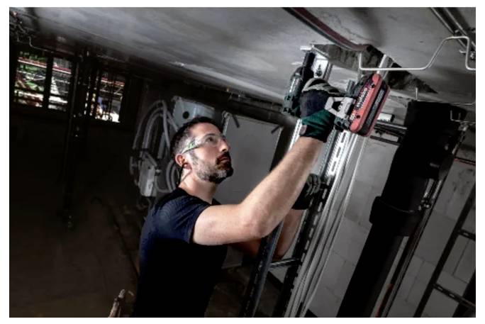 A man wearing safety goggles and gloves uses a power tool while standing on a platform, working on a ceiling structure in an industrial setting.