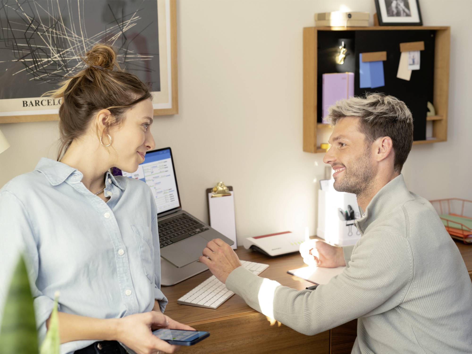 A man and a woman are having a conversation at a desk with a laptop. Office environment with documents and decorations in the background.