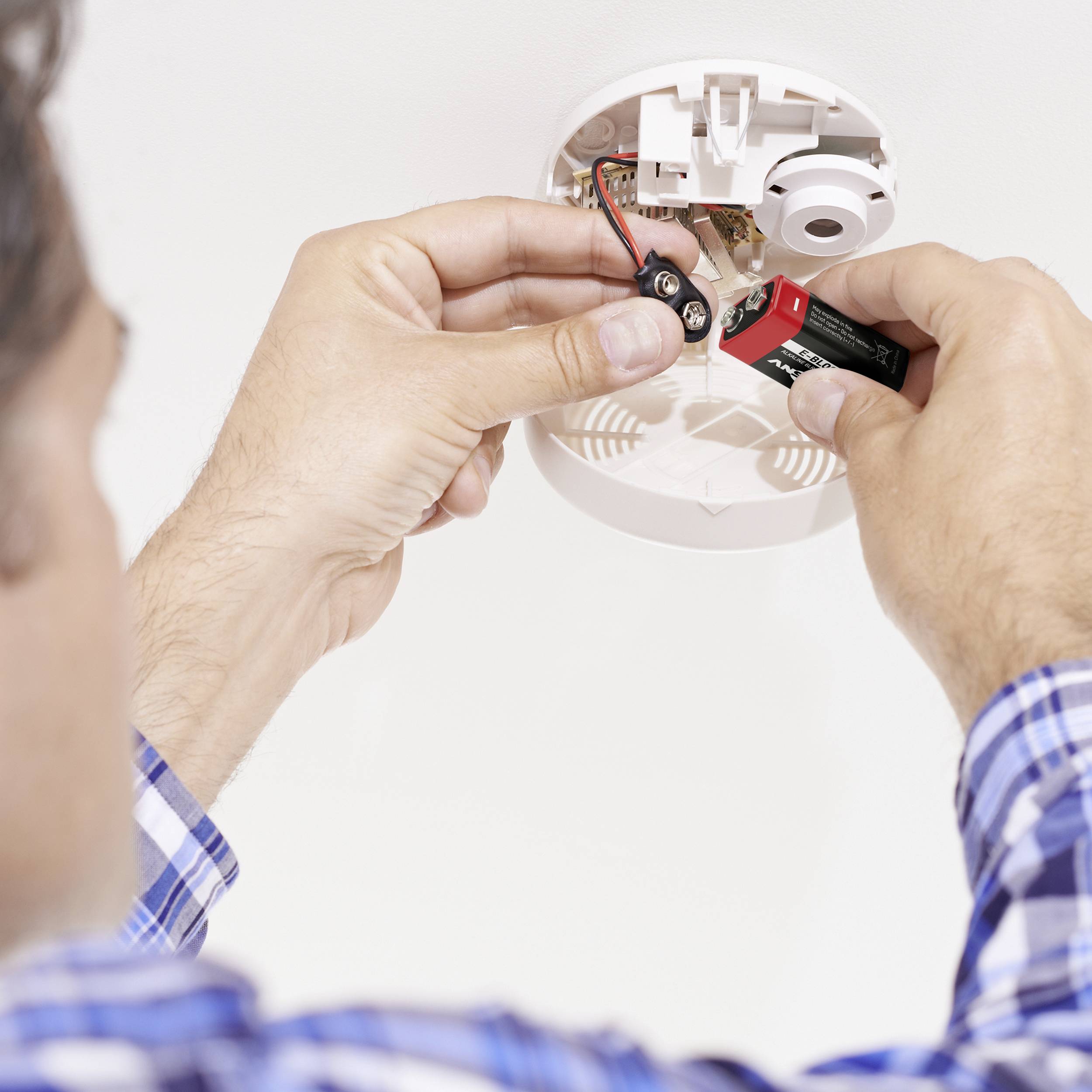 A person is installing a battery in a smoke detector on the ceiling. Hands are visible, with details of the smoke detector in focus.