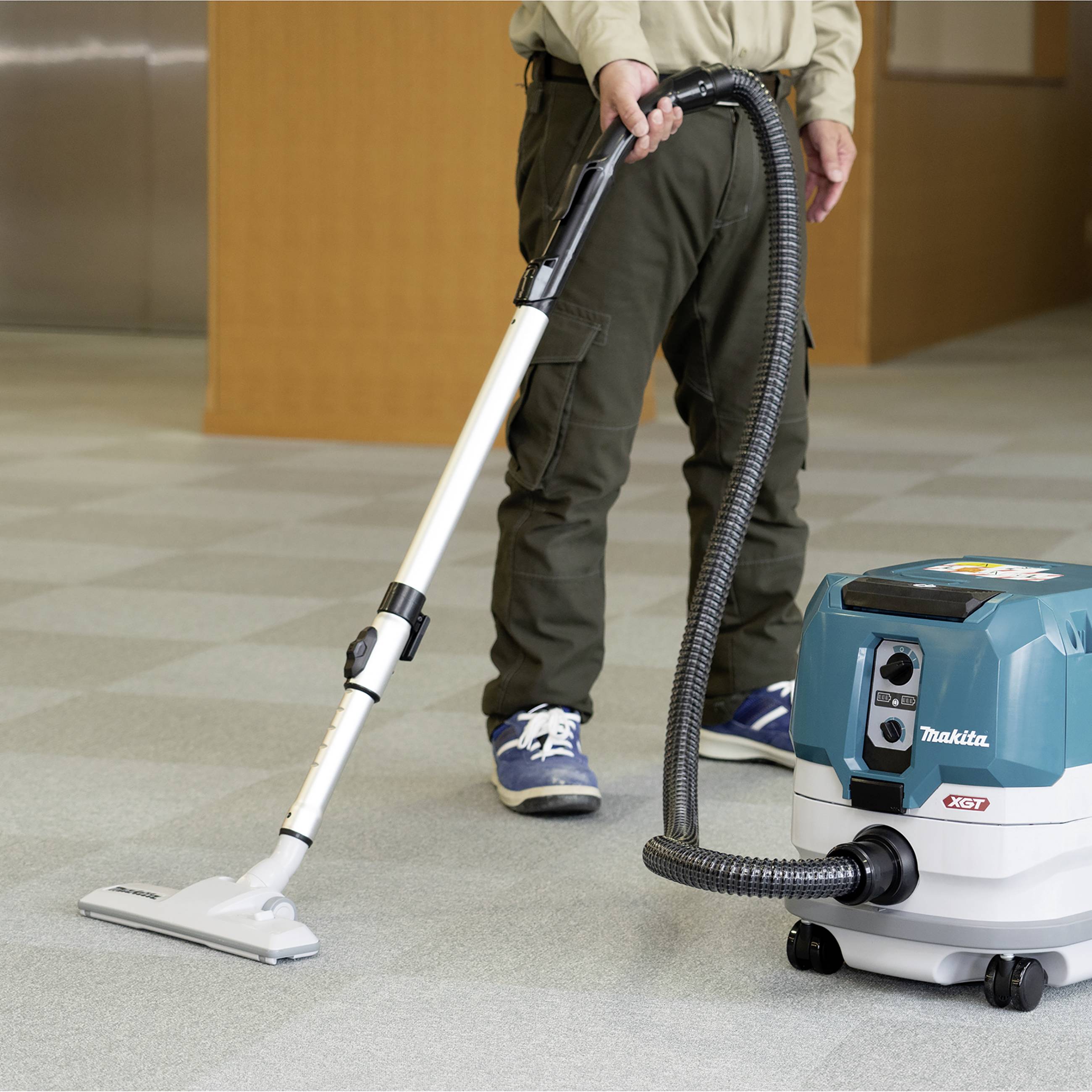 A person is vacuuming the floor with a cordless hoover in a building. The hoover has a collecting container mounted on wheels.