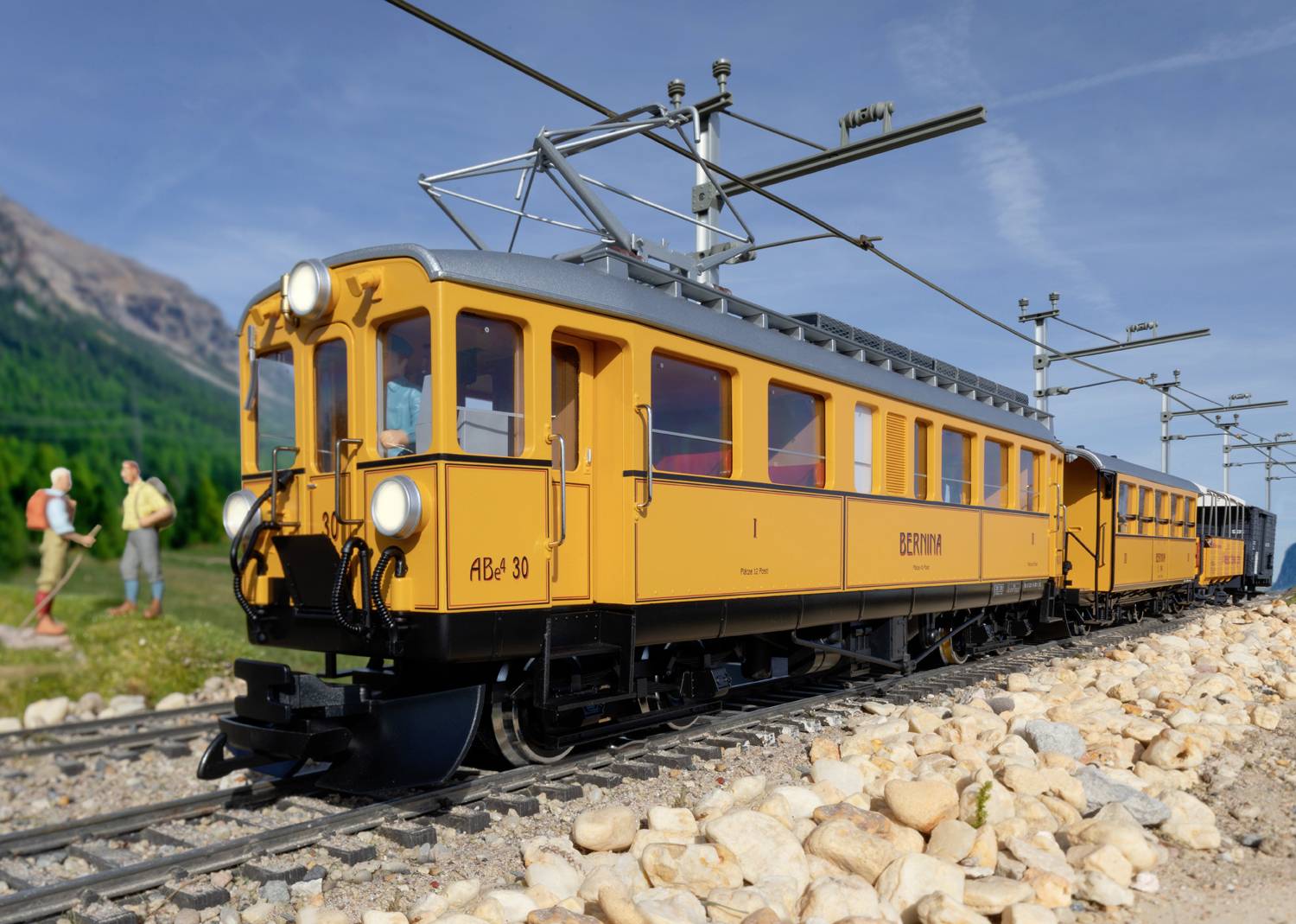 A yellow train travels along a narrow-gauge railway through a rural landscape, with mountains in the background and two people standing by the trackside.