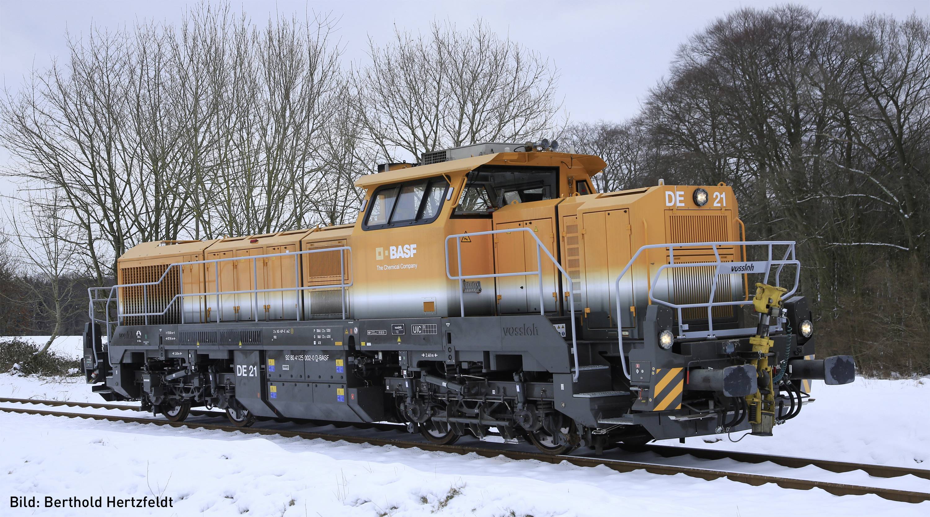 An orange and black locomotive on snow-covered tracks, surrounded by bare trees. A forest is visible in the background.