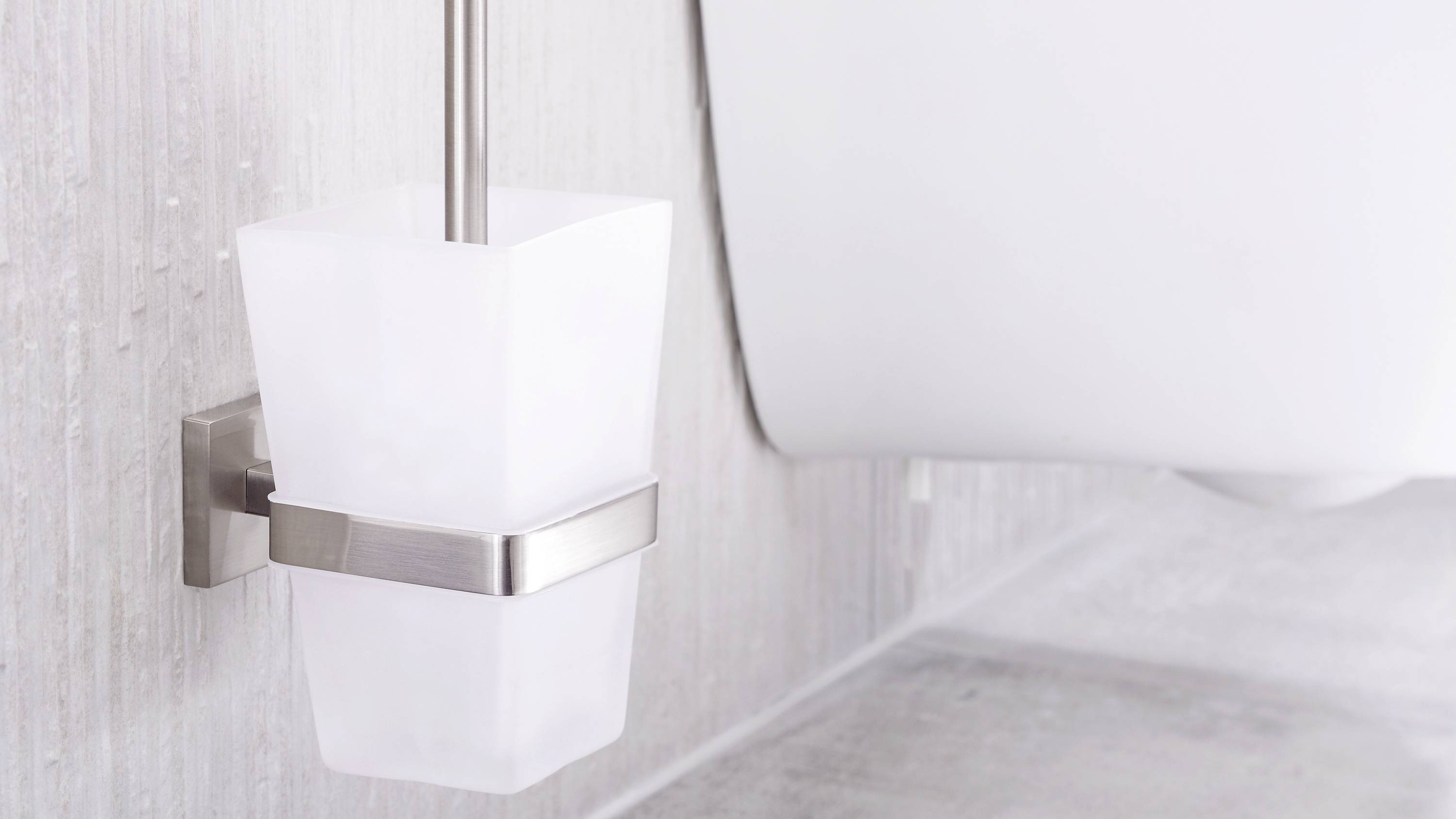 White toilet brush in a modern holder, mounted on a grey wall in the bathroom. Part of the toilet is visible on the right side of the image.