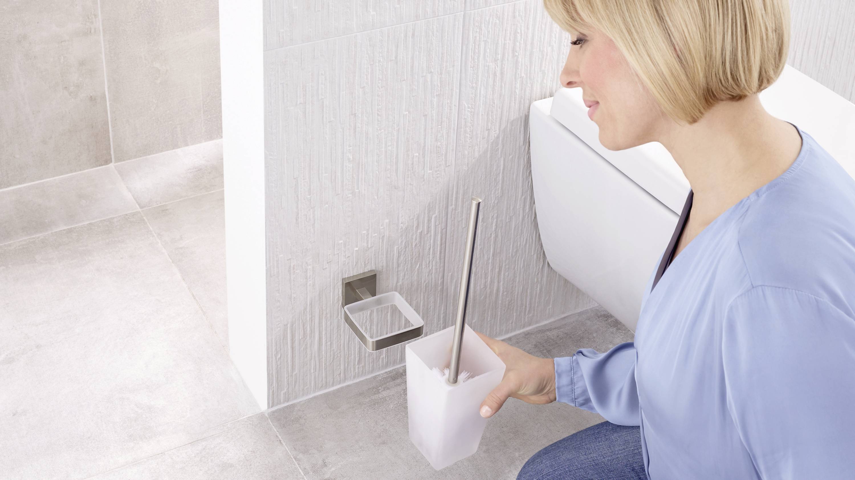 A person is cleaning a bathroom, holding a toilet brush. Tiles and part of the toilet are visible in the background.