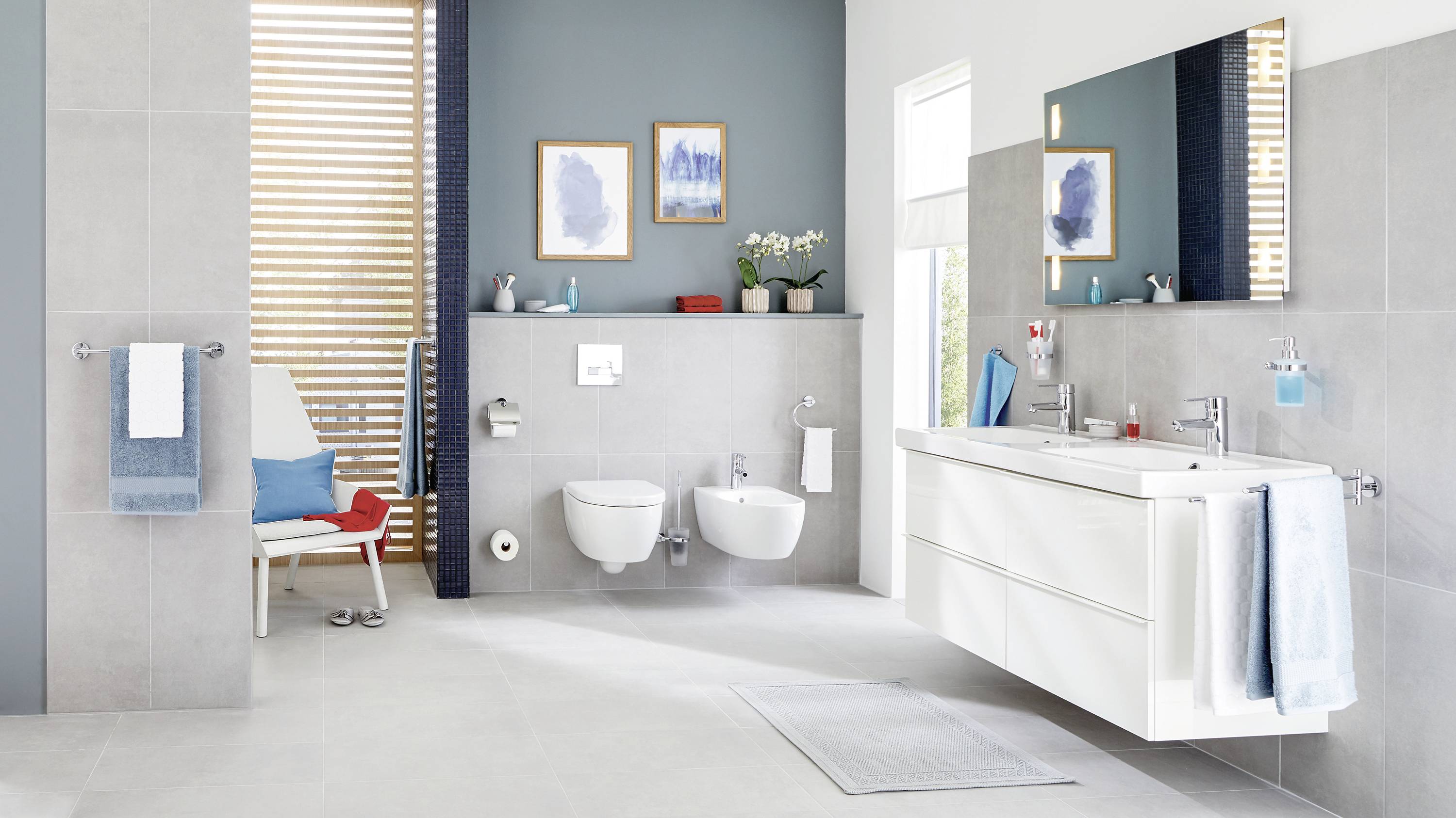 A modern bathroom with grey tiles, wall-mounted toilets, a large mirror, twin washbasins and light blue decorative elements.