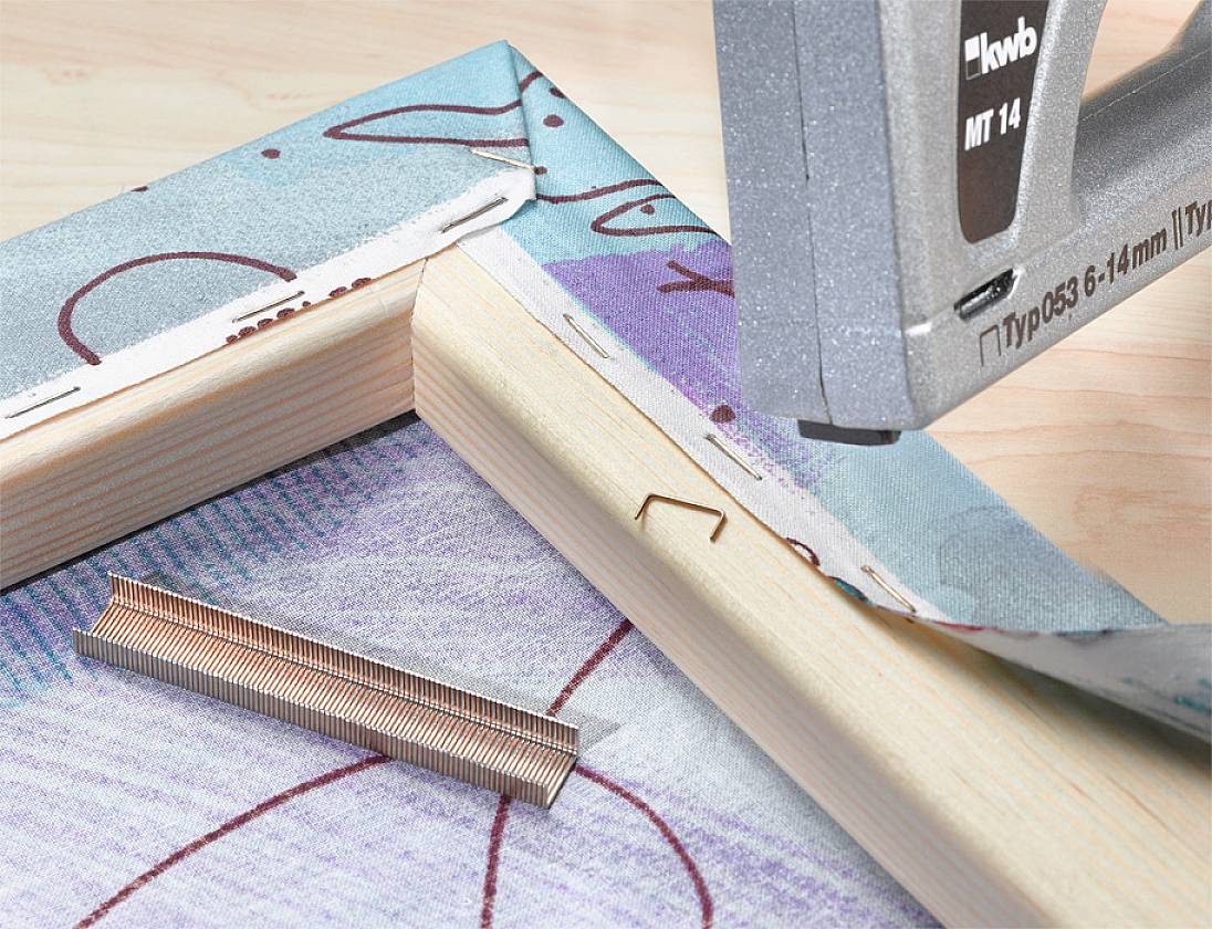 A staple gun fastens canvas to a wooden frame; a strip of staples lies beside it.