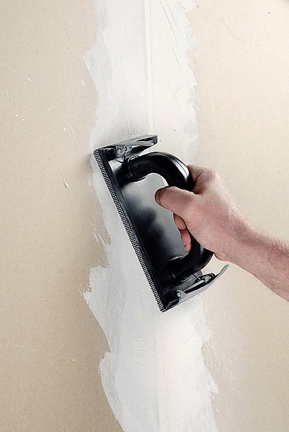 A hand smooths filler with a sanding board on a wall.