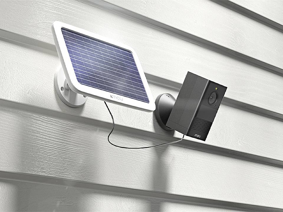 A solar-powered security camera is mounted on a house wall. The solar panel provides power to the camera.