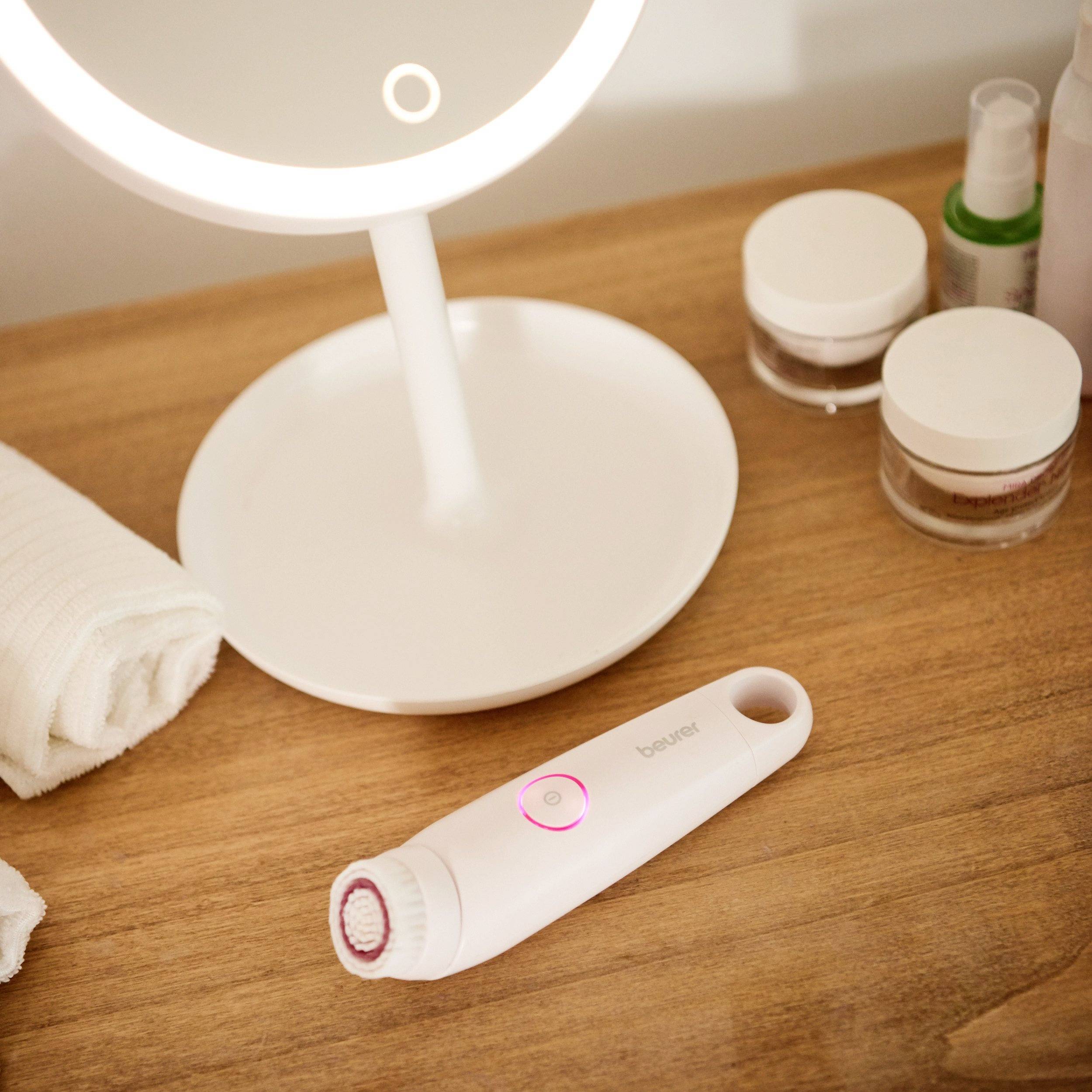 A lit make-up mirror on a wooden table, with an electric cleansing brush beside it. On the table are jars and a cloth.