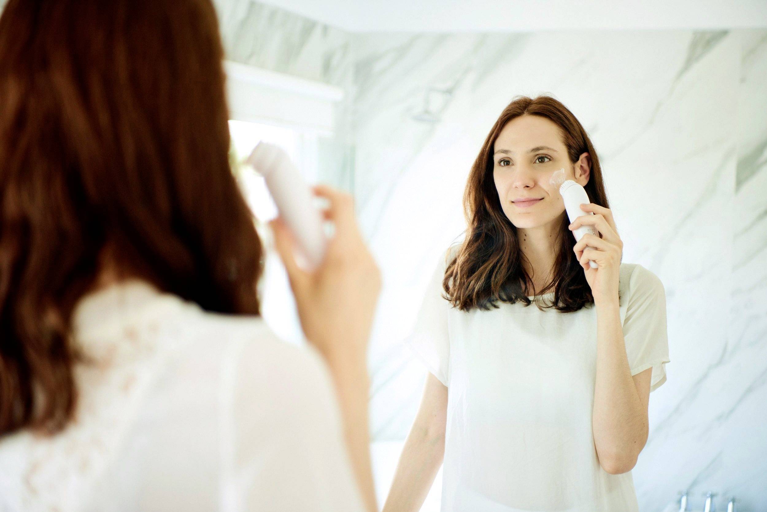 A woman stands in front of a mirror in a bathroom, holding an electric skincare device to her face while examining her reflection.
