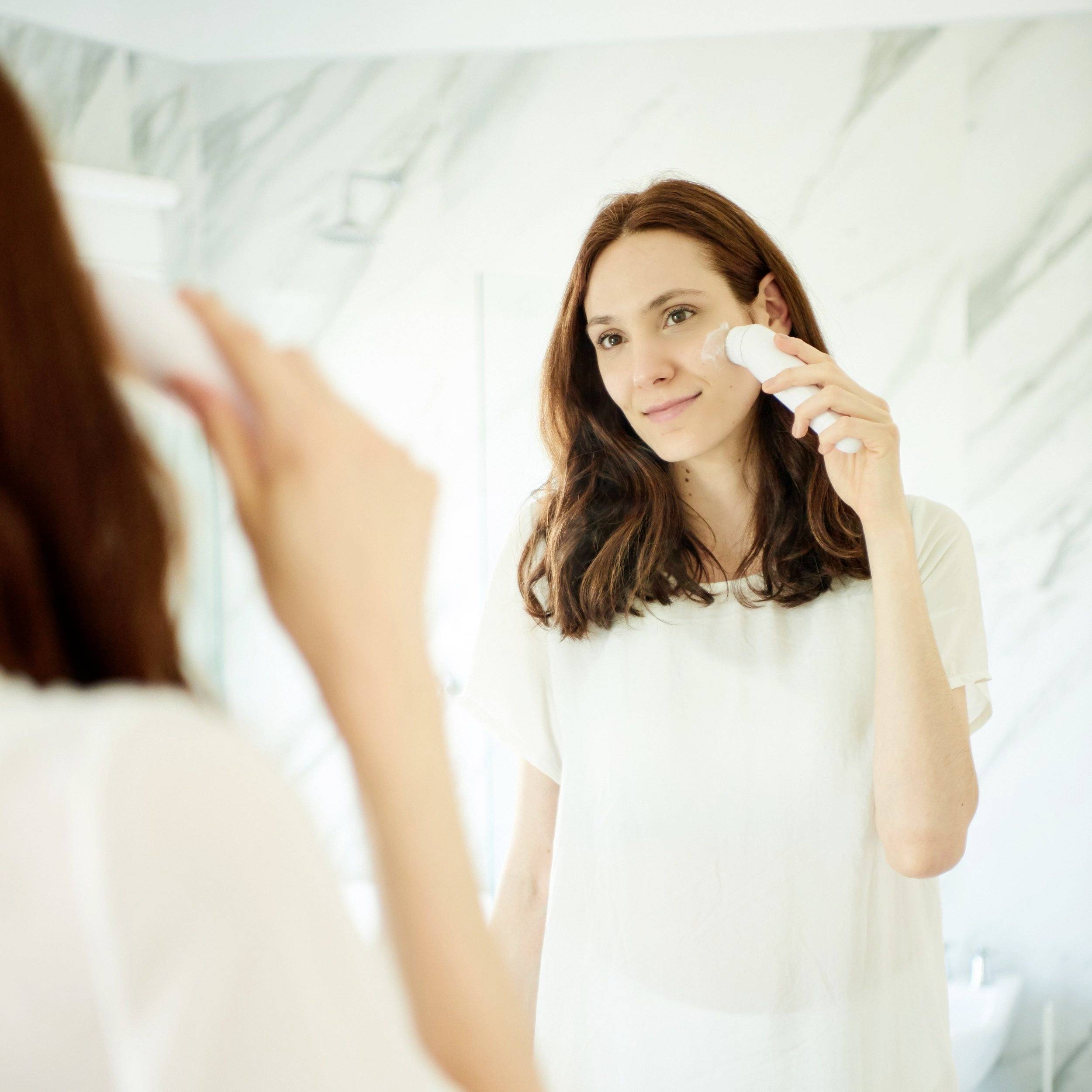 A woman applies a cosmetic product to her face with a cotton pad in a bathroom.