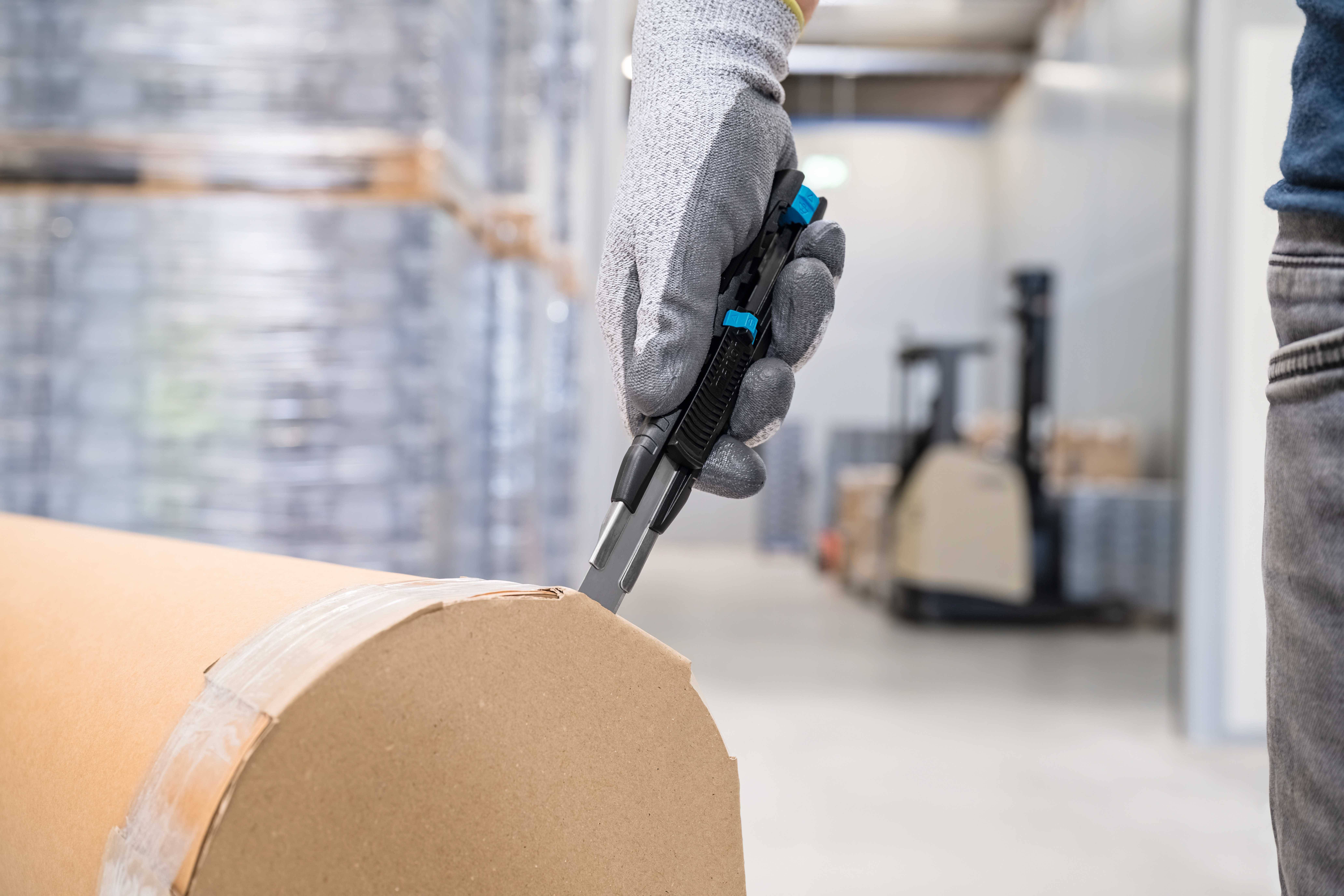 A hand wearing a work glove is holding a utility knife and cutting a cardboard packaging at a large roll in a warehouse.