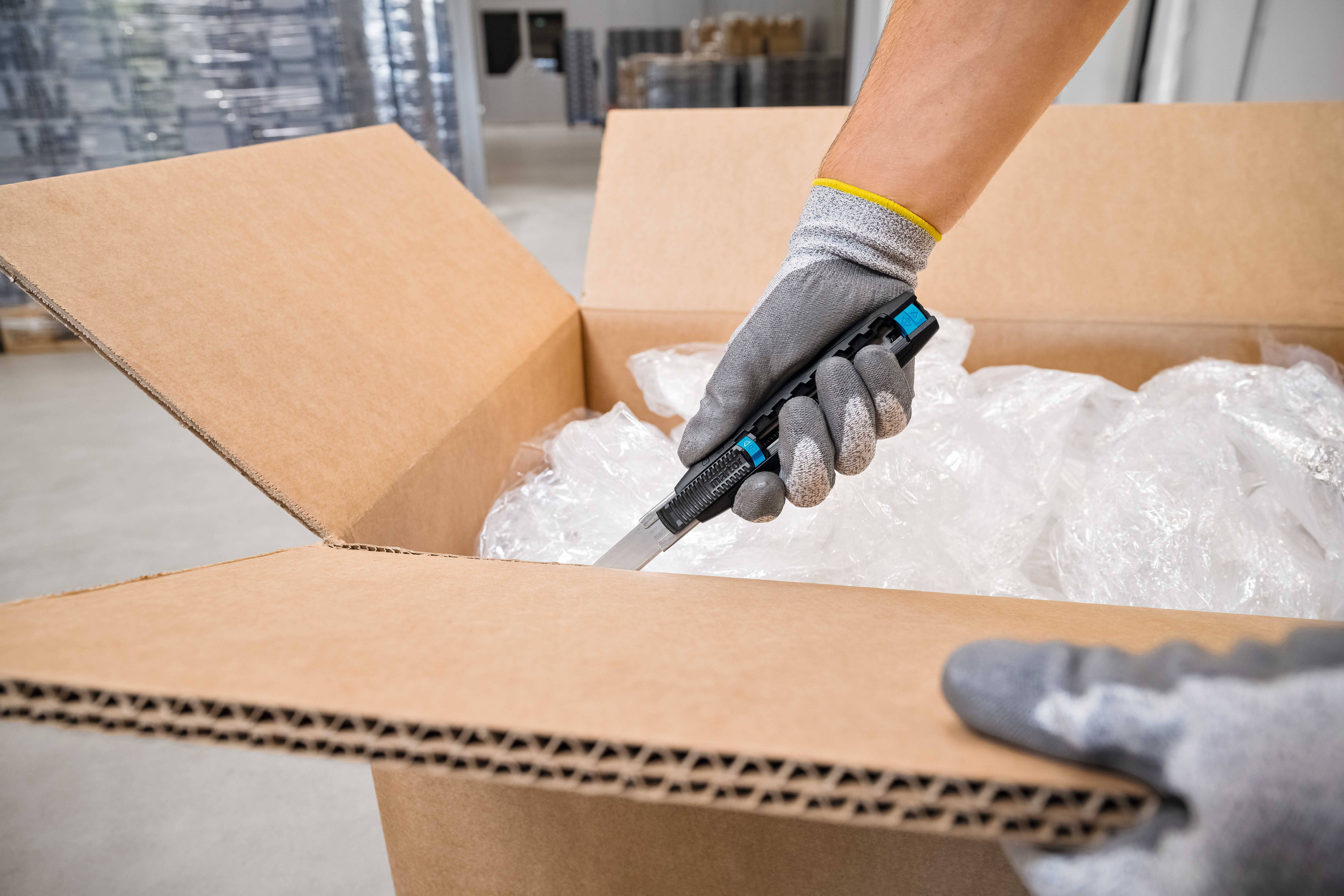 A person opens a cardboard box with a utility knife, which is filled with packaging material. Warehouse shelves are visible in the background.