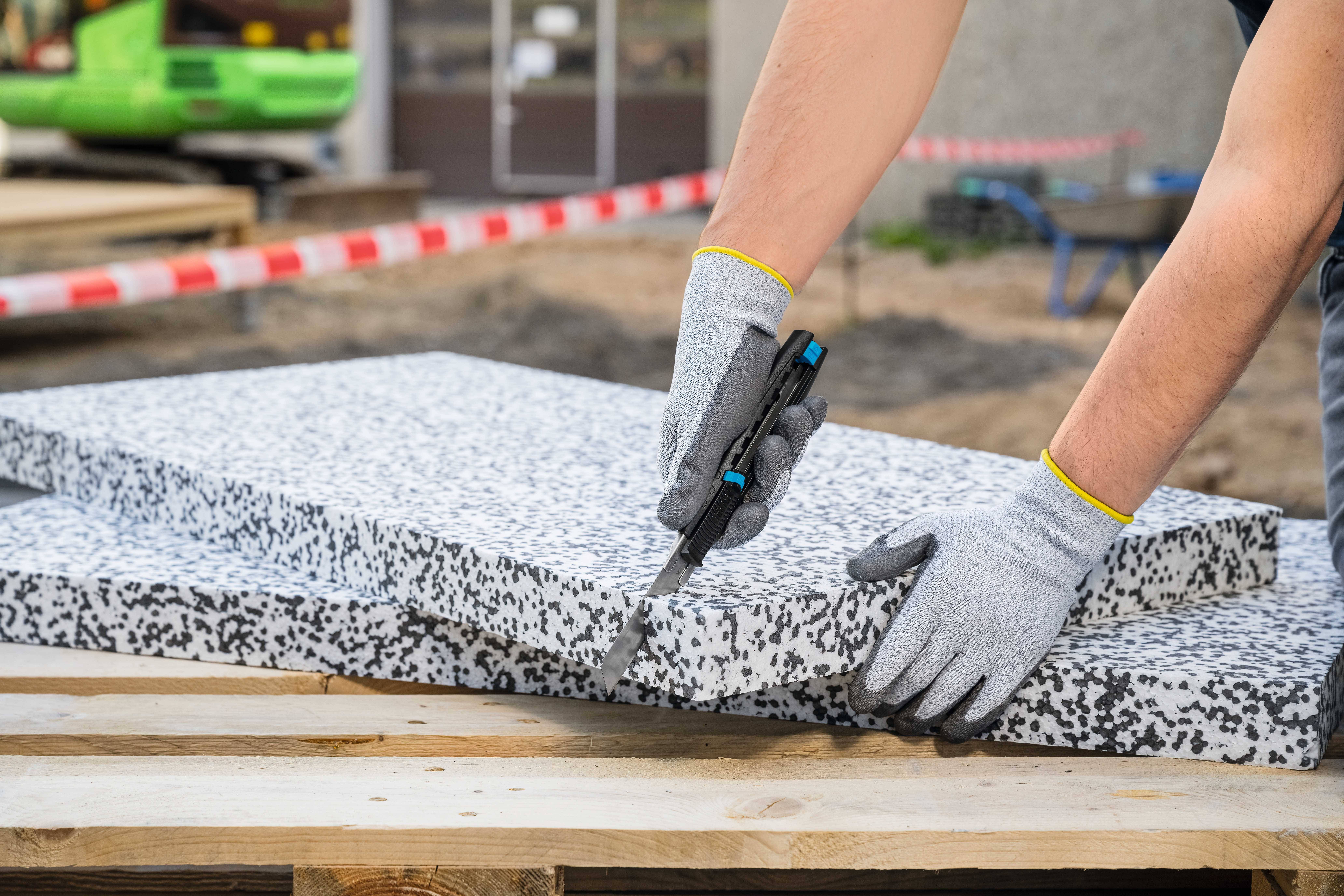 A person is cutting a grey polystyrene sheet with a knife. They are wearing grey gloves. A construction site can be seen in the background.