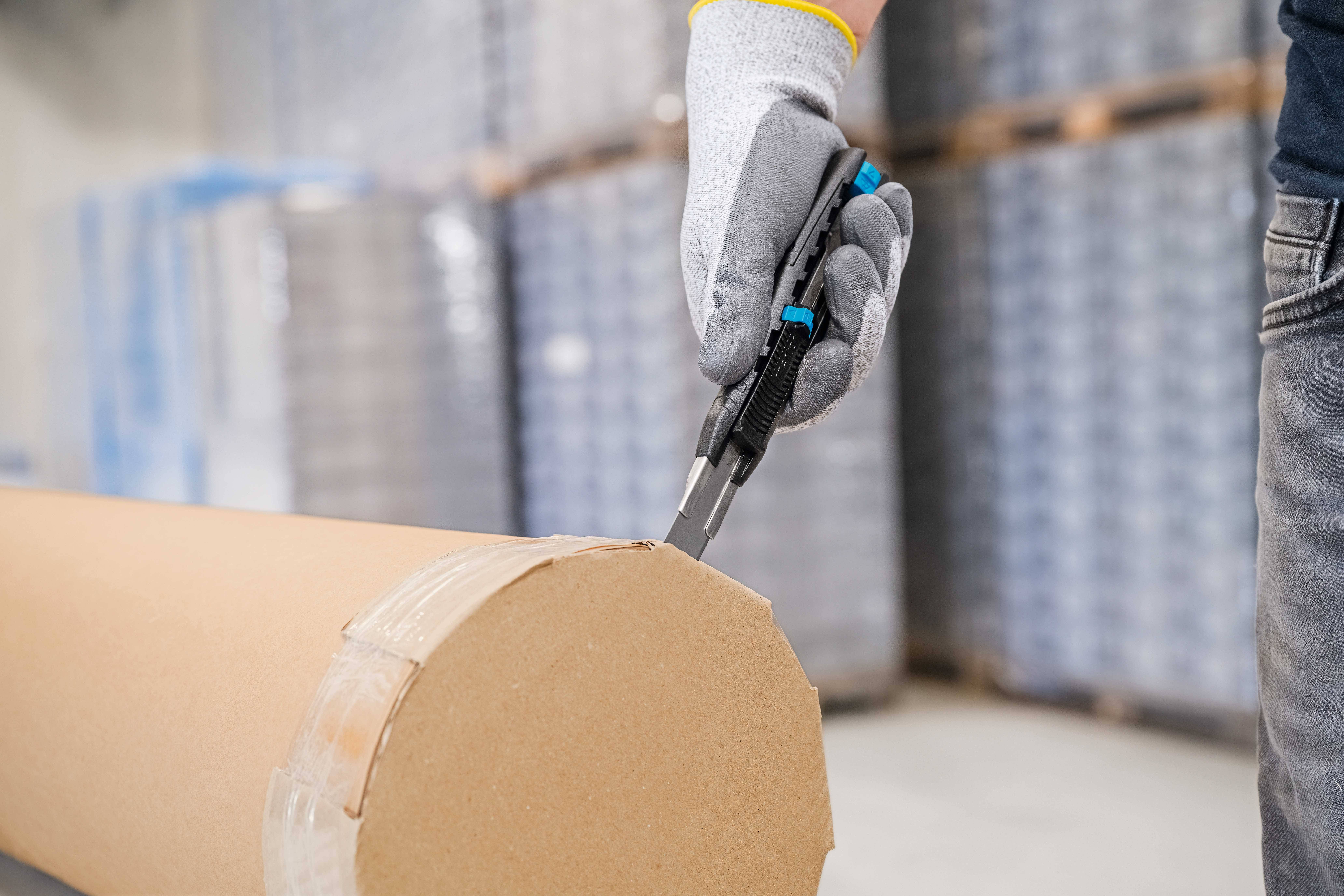 A person is cutting cardboard packaging with a Stanley knife. Background blurred with storage rooms and shelves.