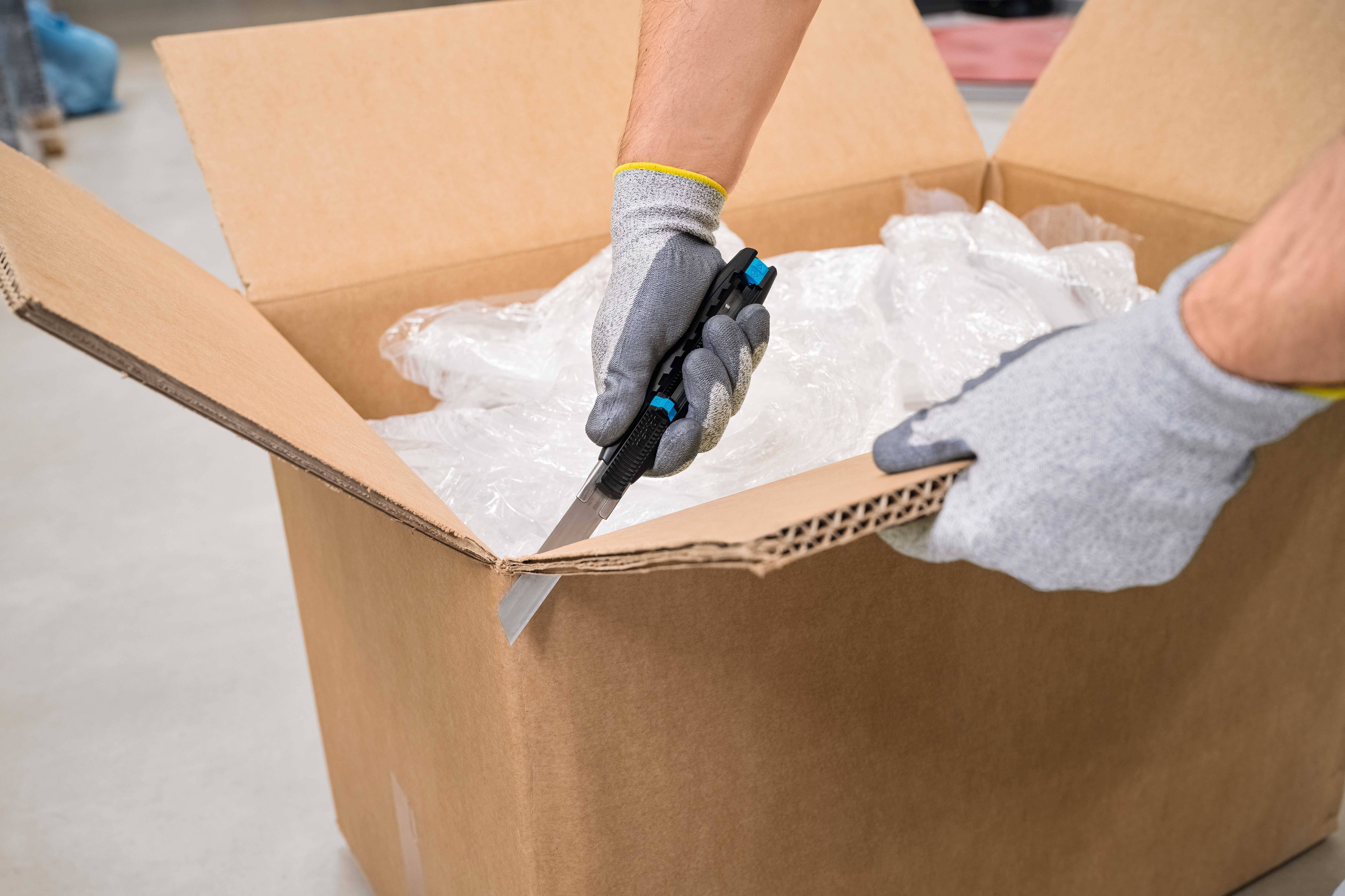 A person is cutting open a cardboard box with a knife. The box contains plastic packaging material. The person is wearing grey gloves.