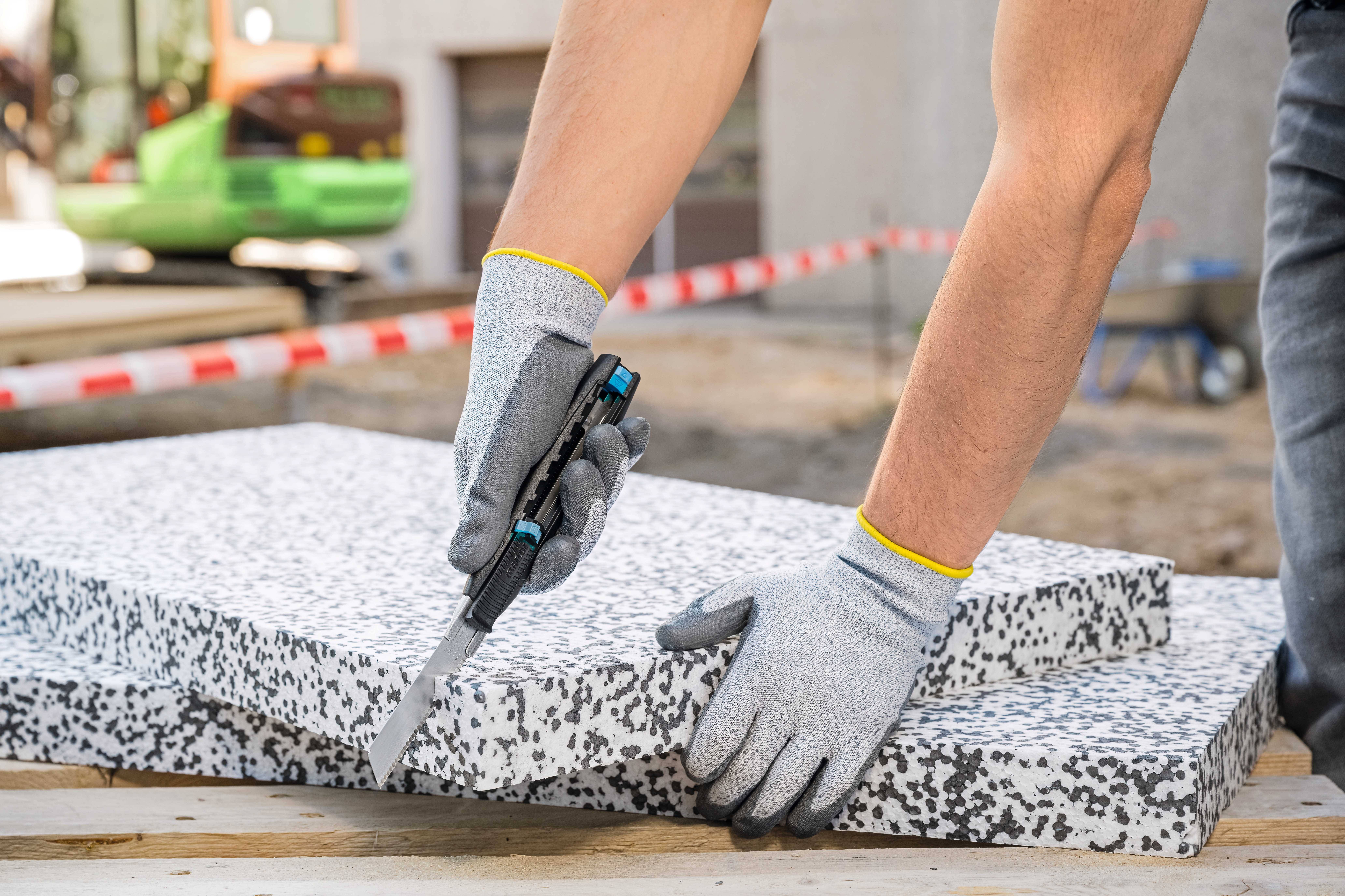 A person wearing grey gloves is cutting a polystyrene board with a knife. A construction site can be seen in the background.