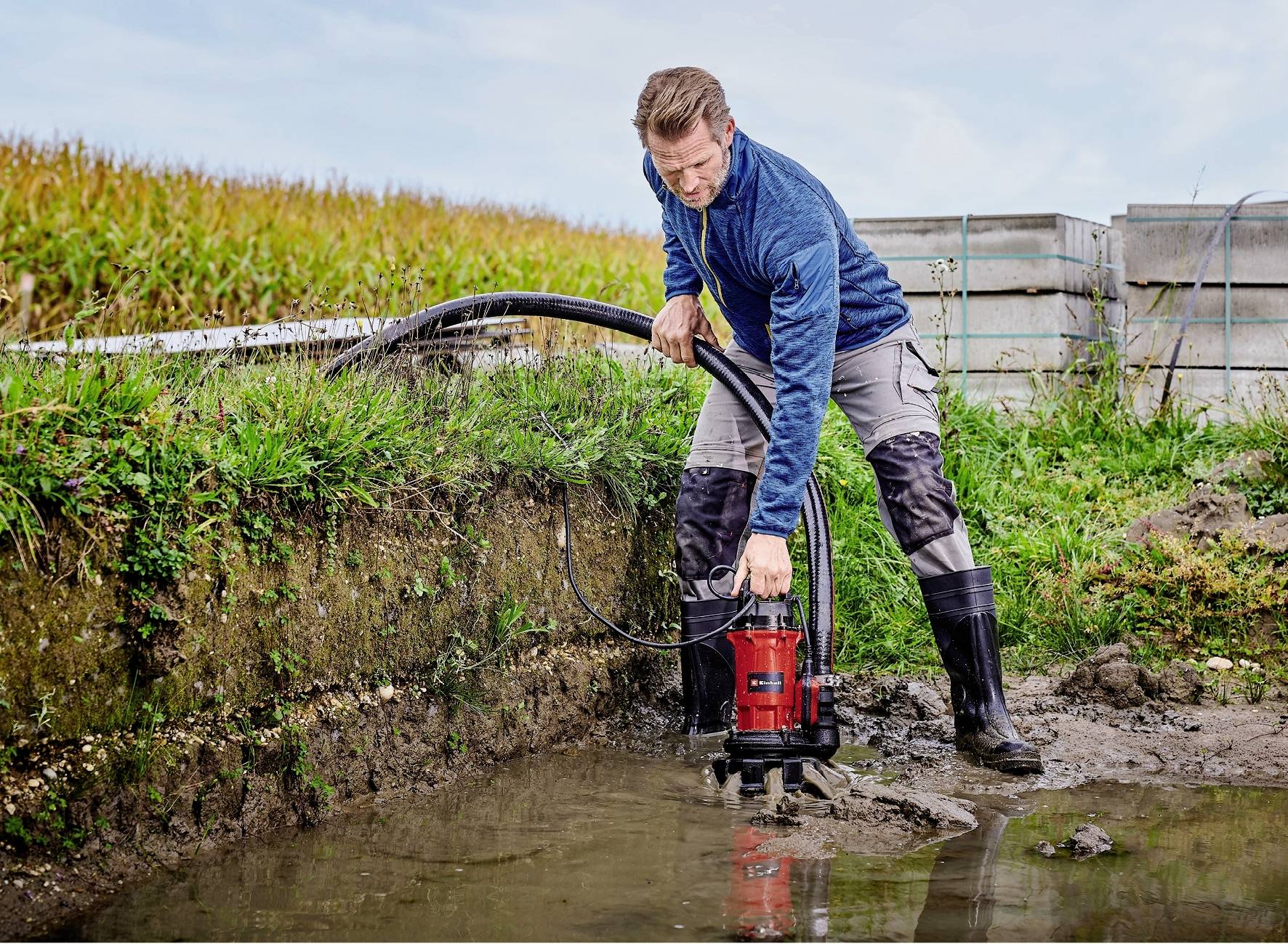 A man in workwear is pumping water from a ditch using a small, red water pump. A field can be seen in the background.