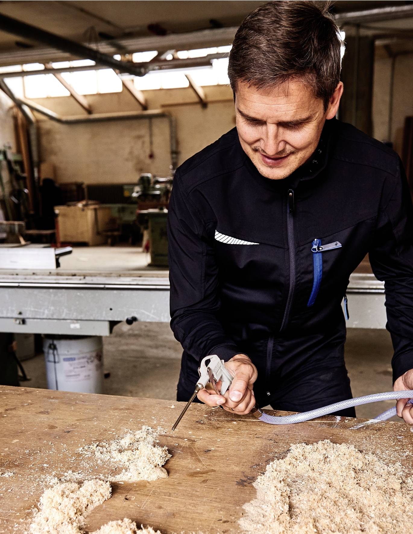 A man in a workshop is cutting wood with a saw on a workbench. Tools and machinery can be seen in the background.