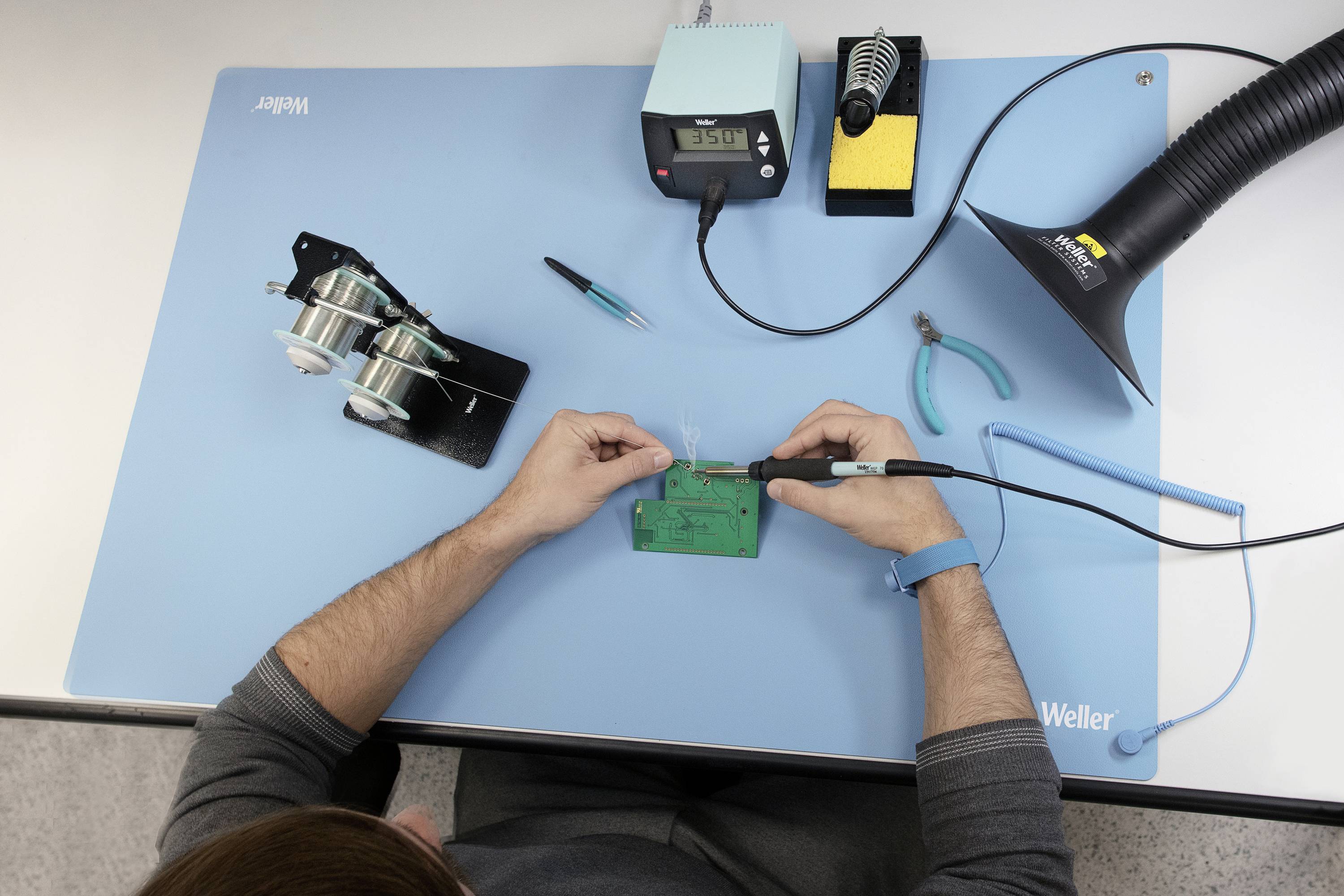 A person is soldering on a circuit board on a blue mat. Next to the person is soldering equipment, including a soldering iron stand, solder wires, and an extraction system.