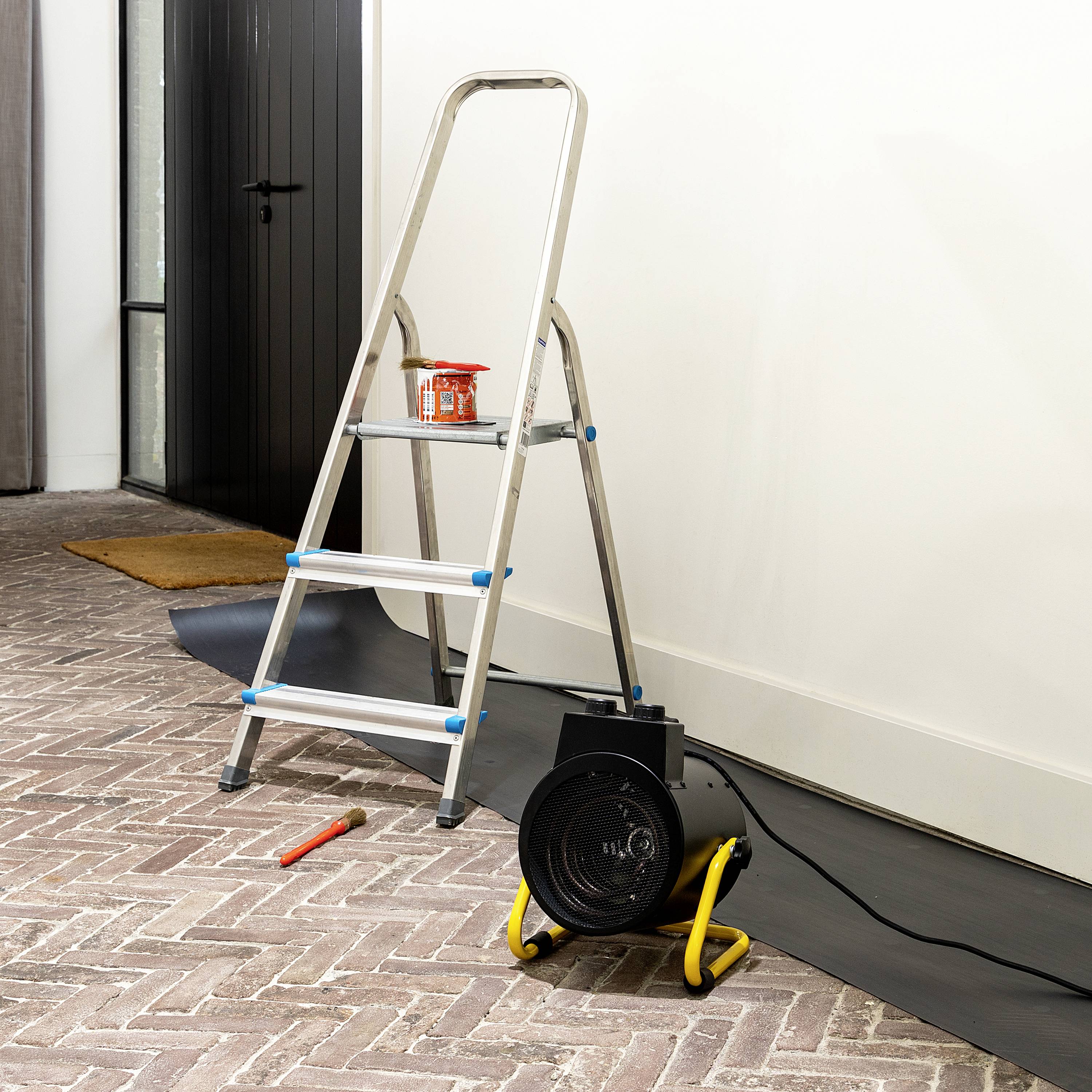 A ladder is standing on a tiled floor next to a wall that is being renovated. A paint bucket with a paintbrush sits on the ladder.