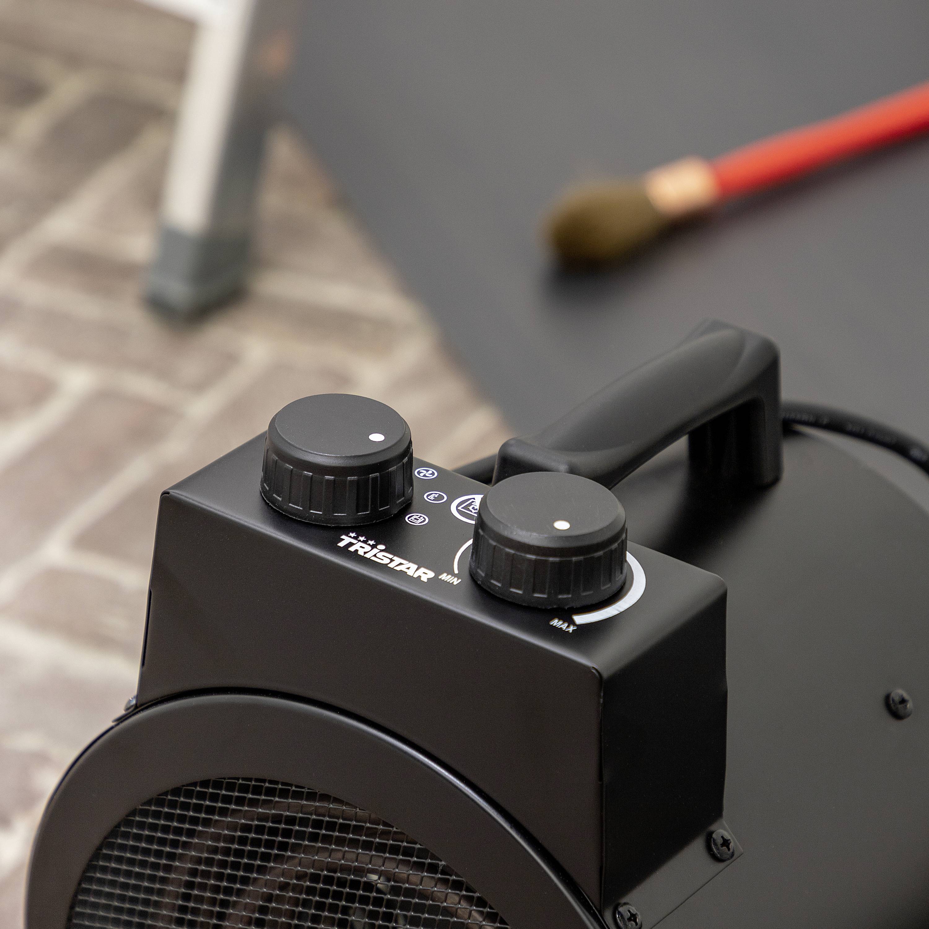 A heating appliance stands on a brick floor surface, with a red broom lying in the background. The focus is on the device's control element.