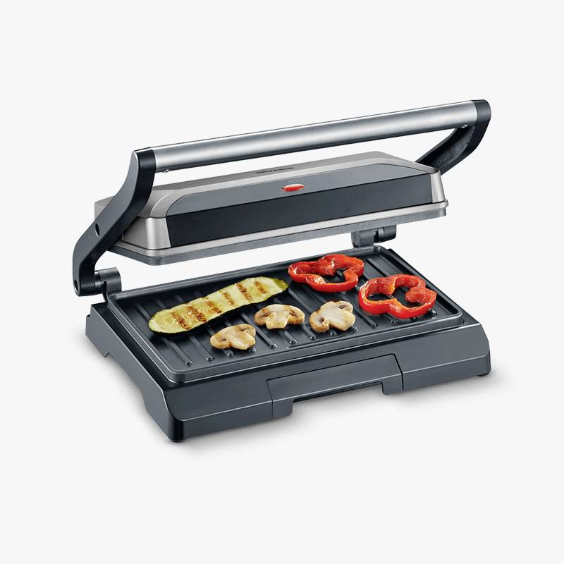 An open tabletop grill with grilled vegetables, including courgettes, peppers and mushrooms, on the ridged grilling surface.