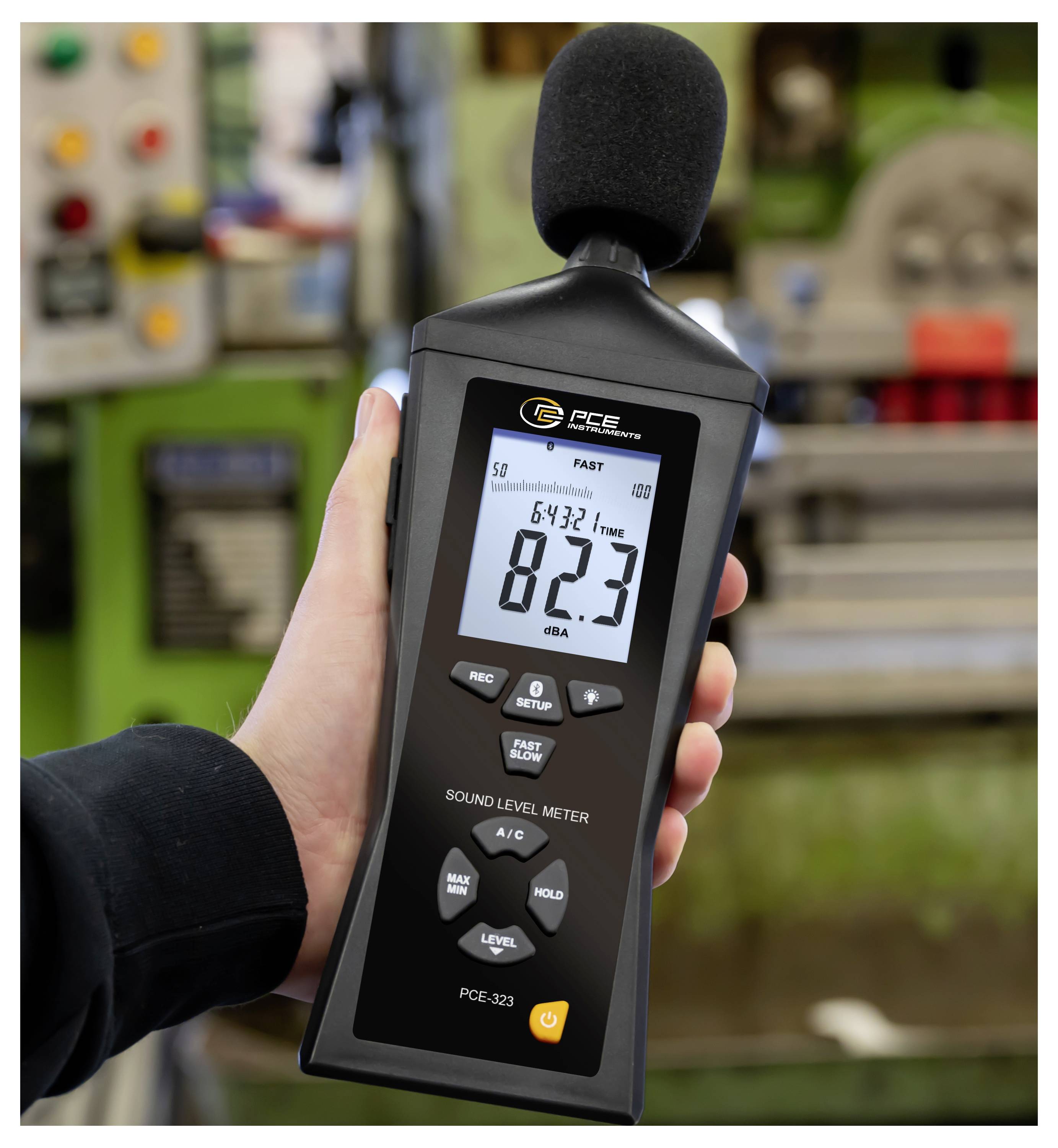 Handheld sound level meter with foam microphone shows 82.3 dBA in a noisy industrial setting.