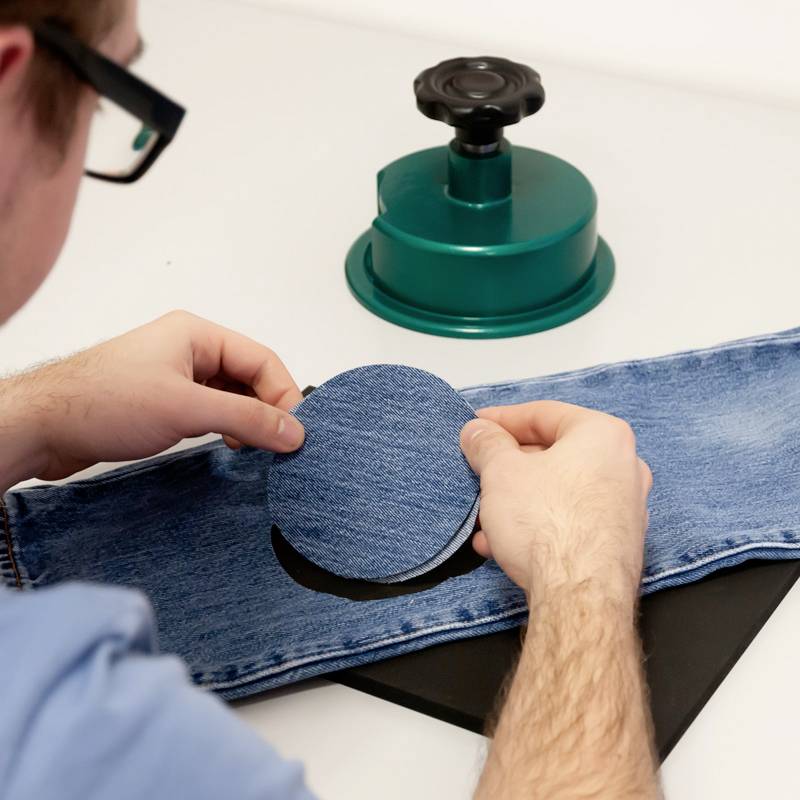 A person is holding a round, blue fabric patch over a pair of jeans, with a green iron nearby.