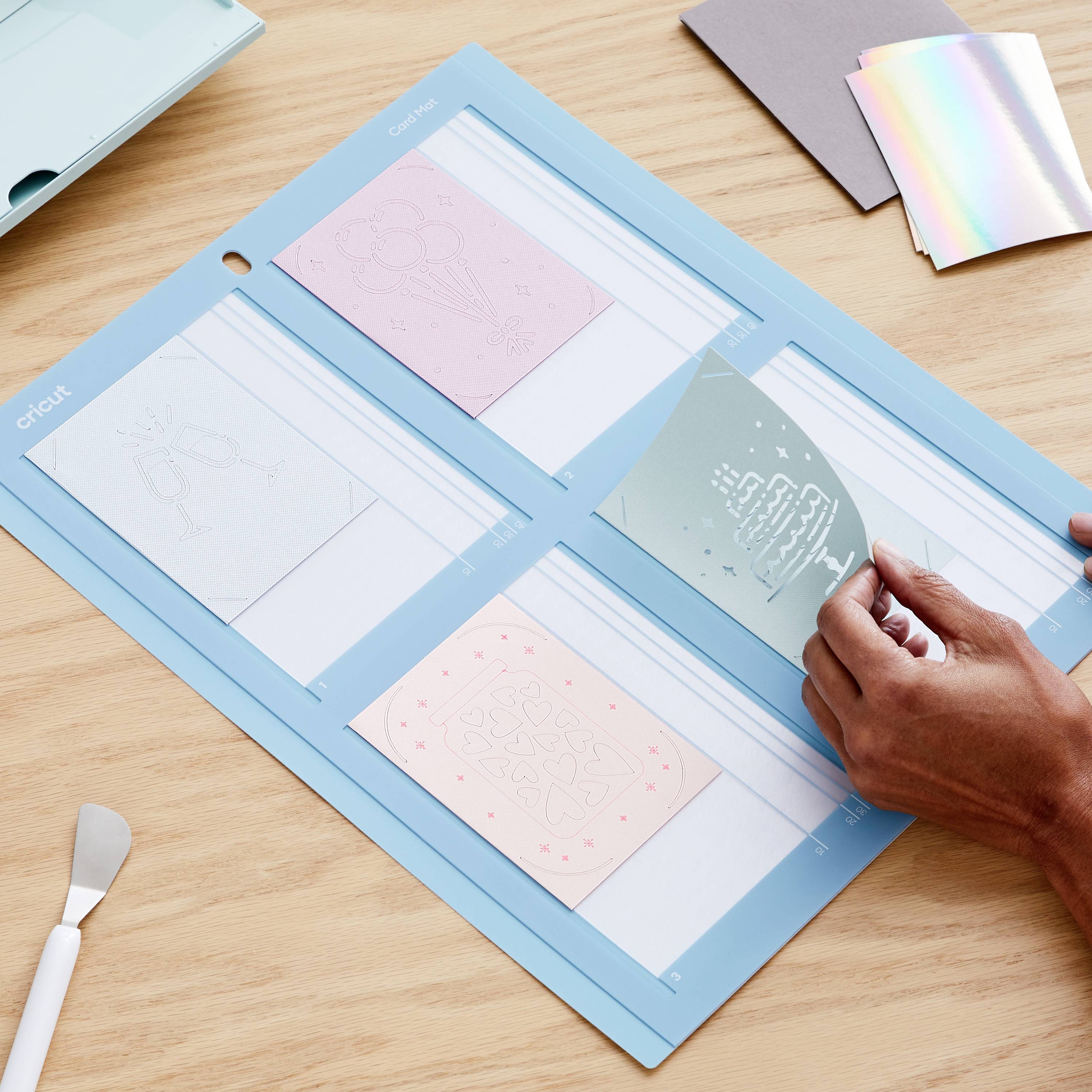 A person is using a Cricut machine to position designs on a blue mat. Various materials and tools are laid out ready.