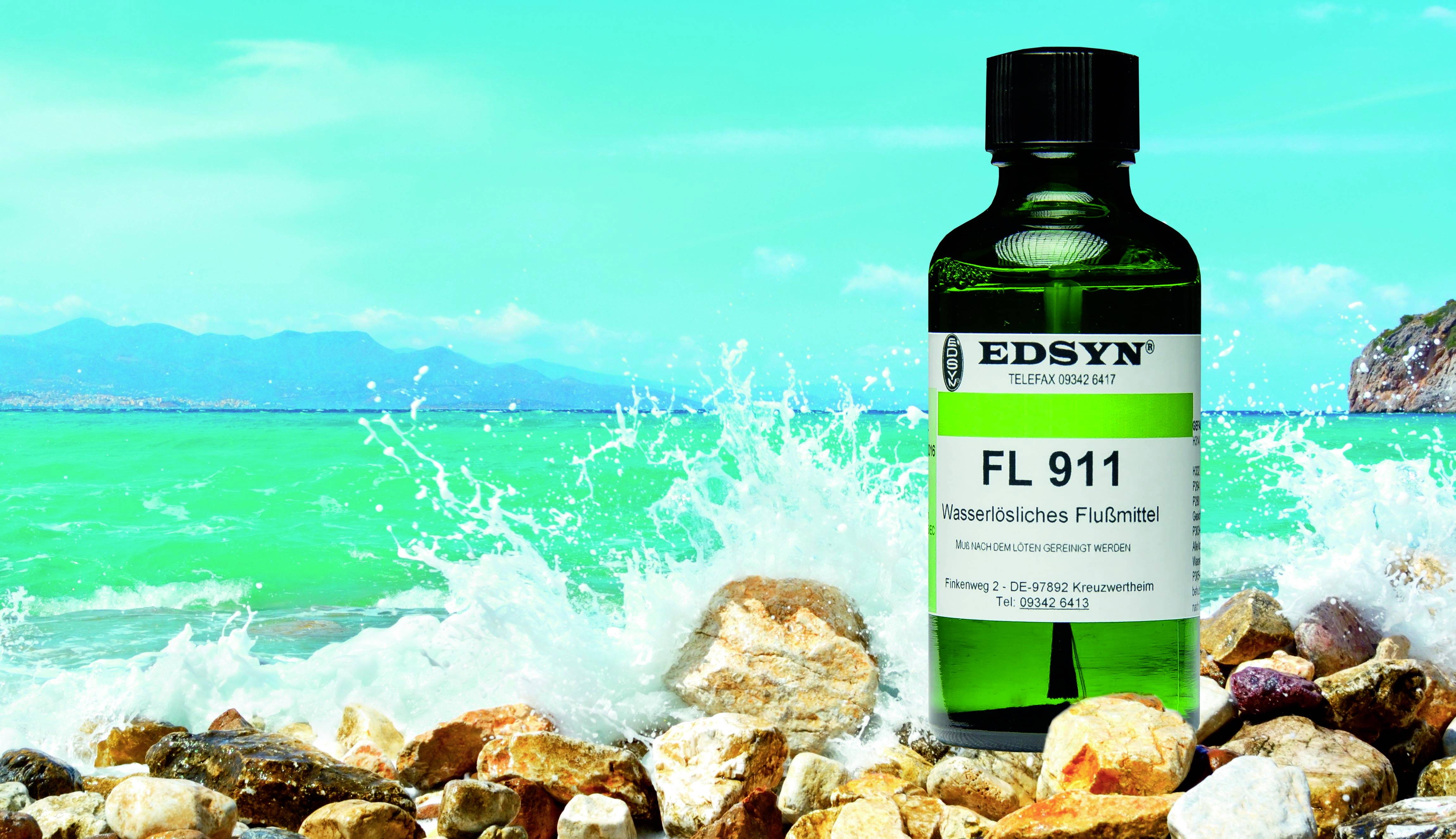 A bottle of 'EDSYN FL 911' water-soluble flux sits on rocks by the sea. Waves and mountains are visible in the background.