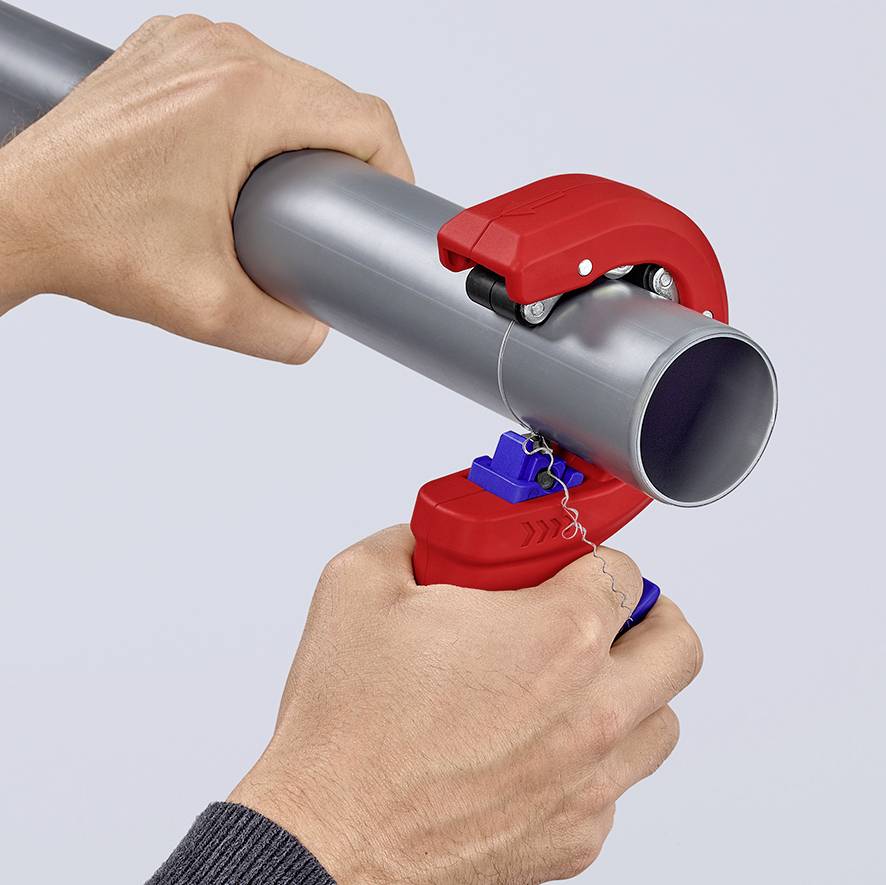 A person is cutting a grey pipe with a red pipe cutter. Hands are holding the cutter around the pipe, small metal shavings are falling off.