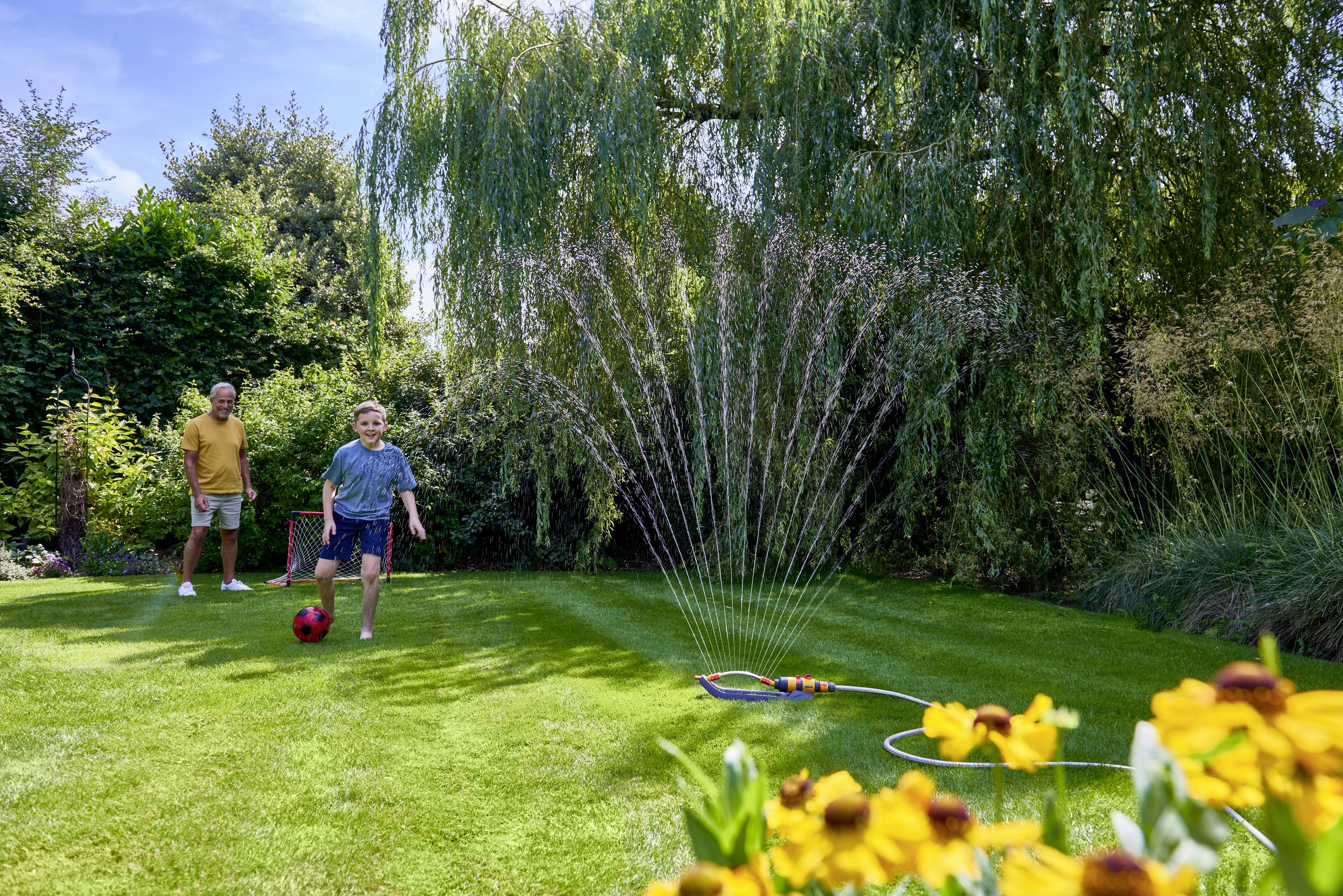 A boy is playing with a red ball in the garden, while an older man watches. A lawn sprinkler is watering the grass. Flowers in the foreground.