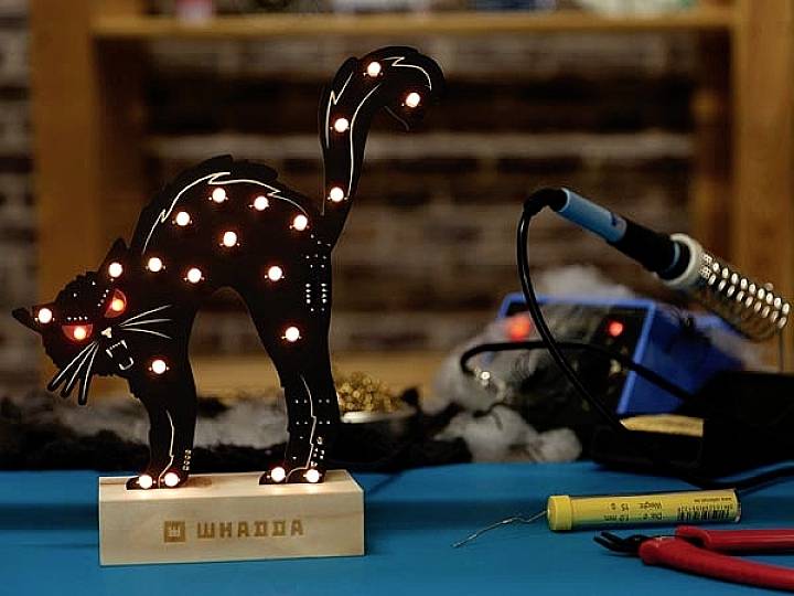 A black cat with glowing LEDs stands on a table next to a soldering iron and electronic tools.